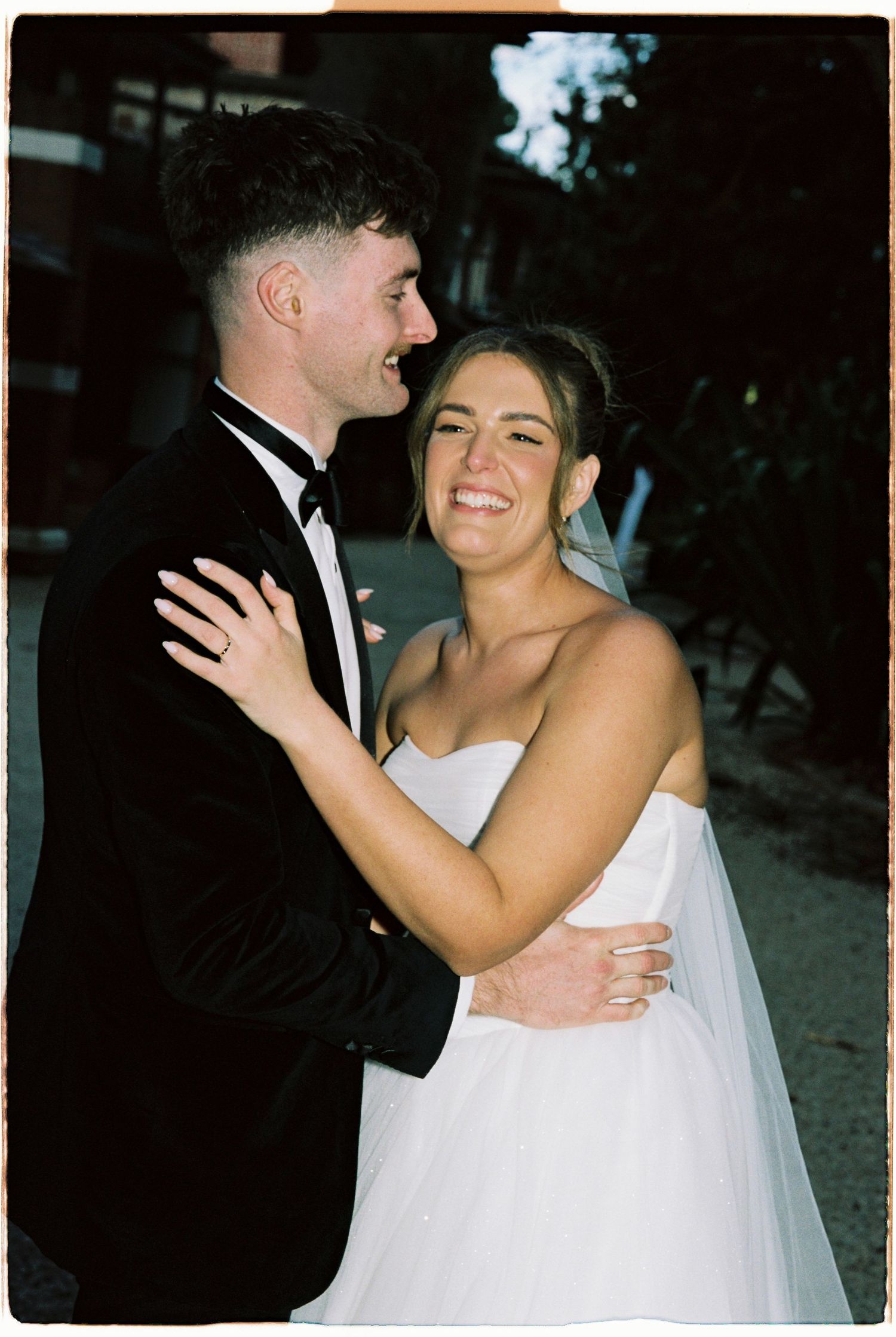 Bride in white strapless gown smiles joyfully as groom in black tuxedo embraces her outdoors at wedding.