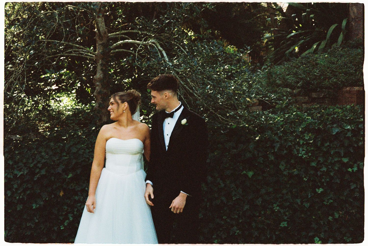Bride in white strapless gown and groom in black tuxedo pose together in lush garden setting.