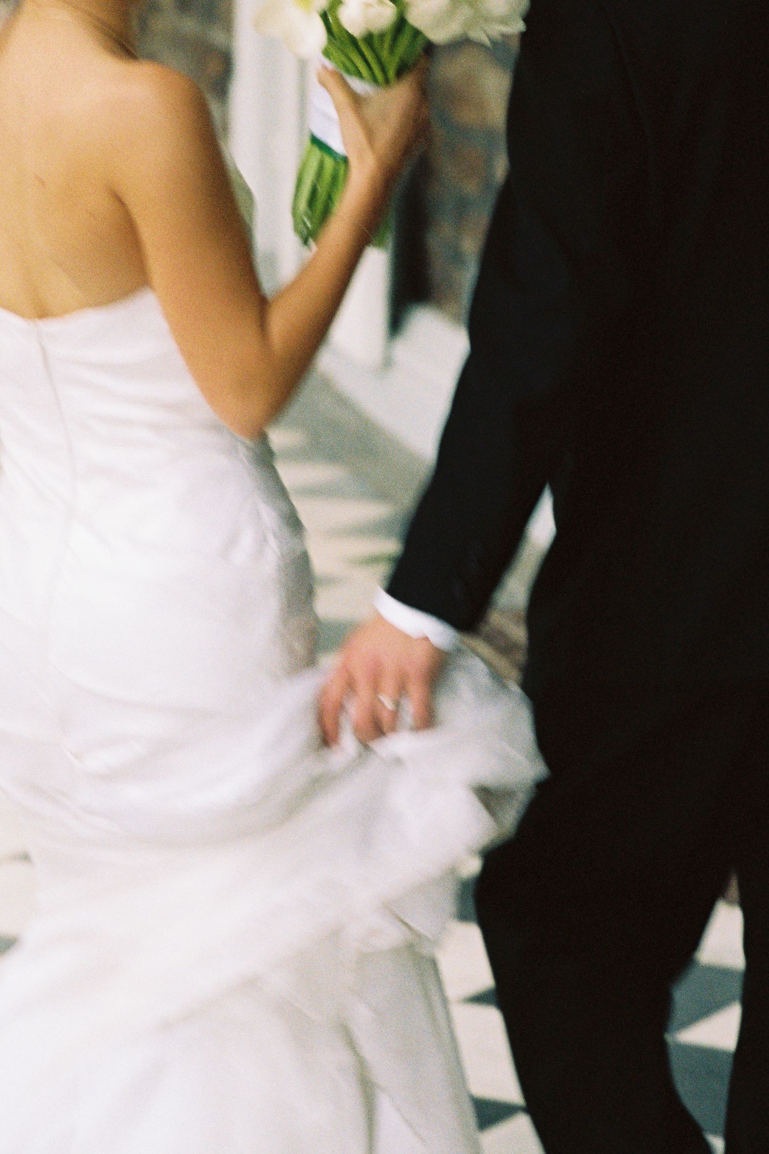 Bride in white strapless gown holding tulip bouquet, holding hands with groom in black suit at wedding ceremony.