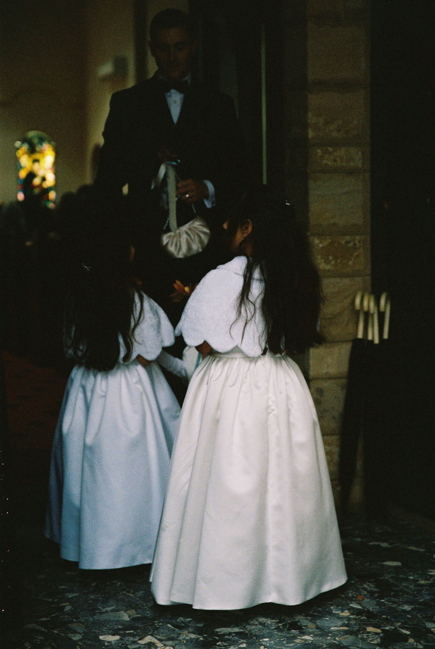 Two young flower girls in white dresses stand near church entrance with a man in formal attire during wedding.