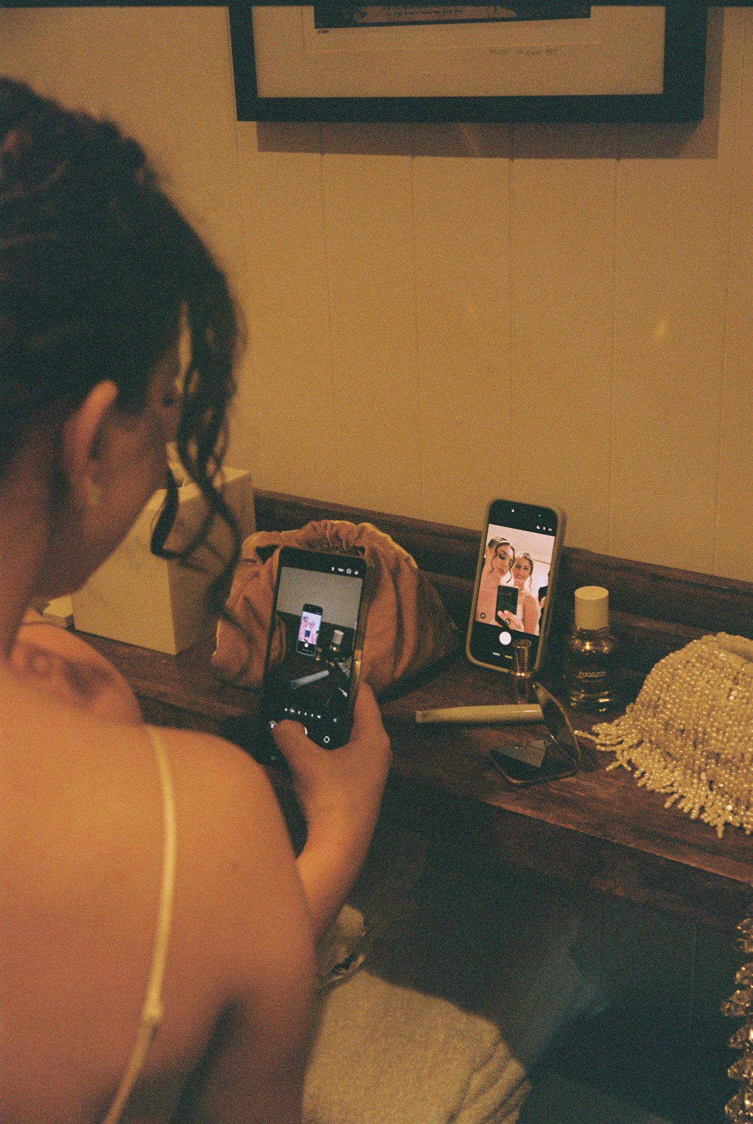 Woman in ivory dress photographs herself using two smartphones propped on wooden vanity table.