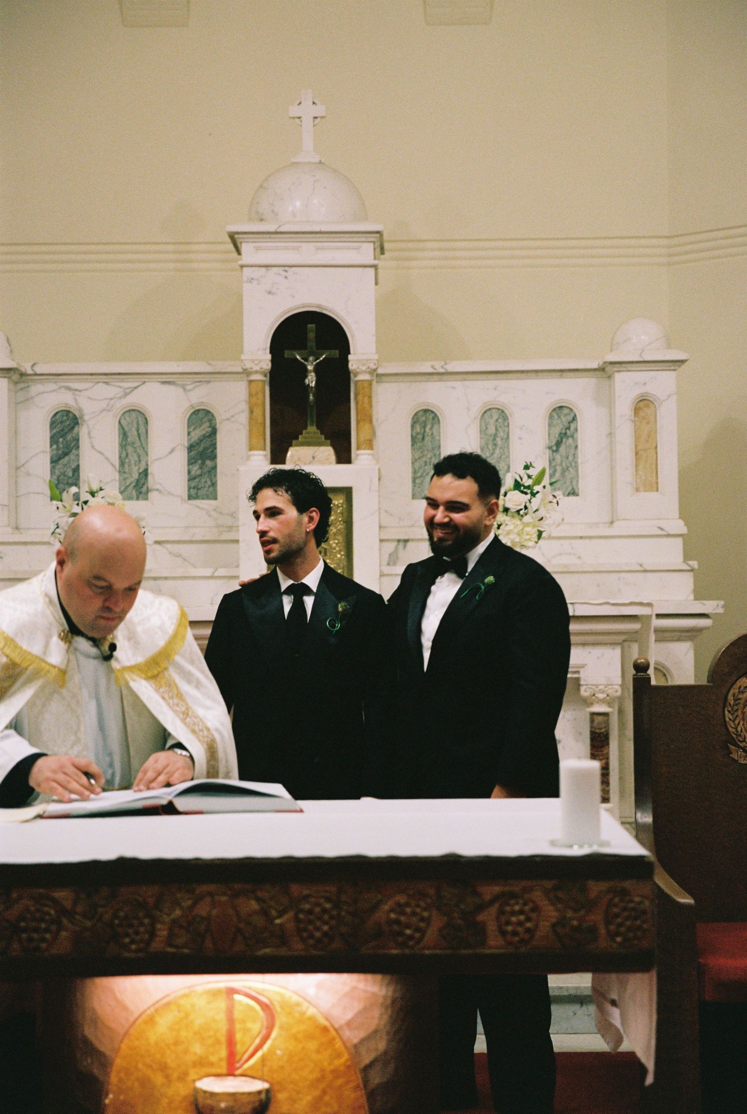 Priest signs wedding register at ornate church altar flanked by groom and best man.