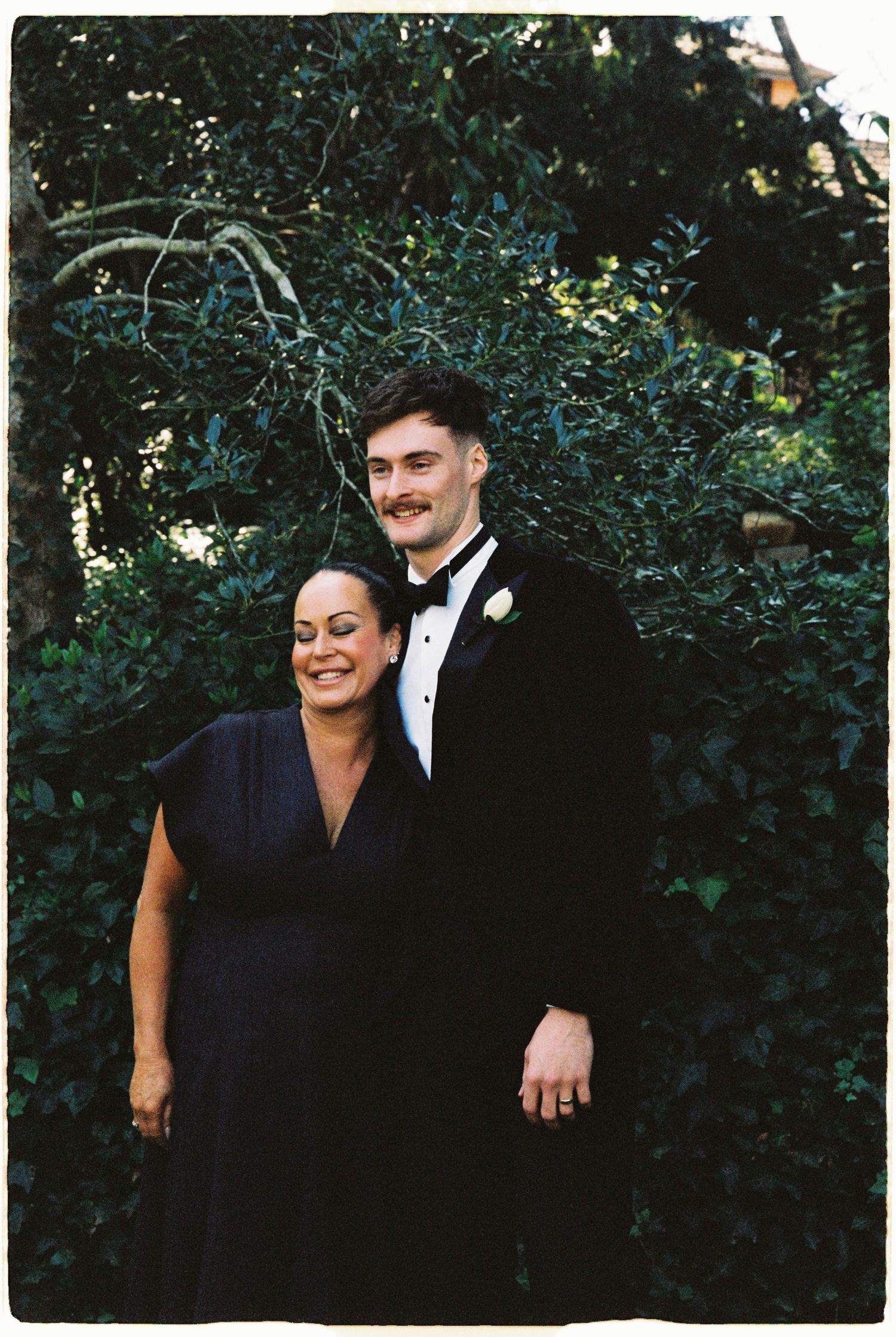 Smiling groom in black tuxedo poses with woman in navy dress amid green foliage at wedding.