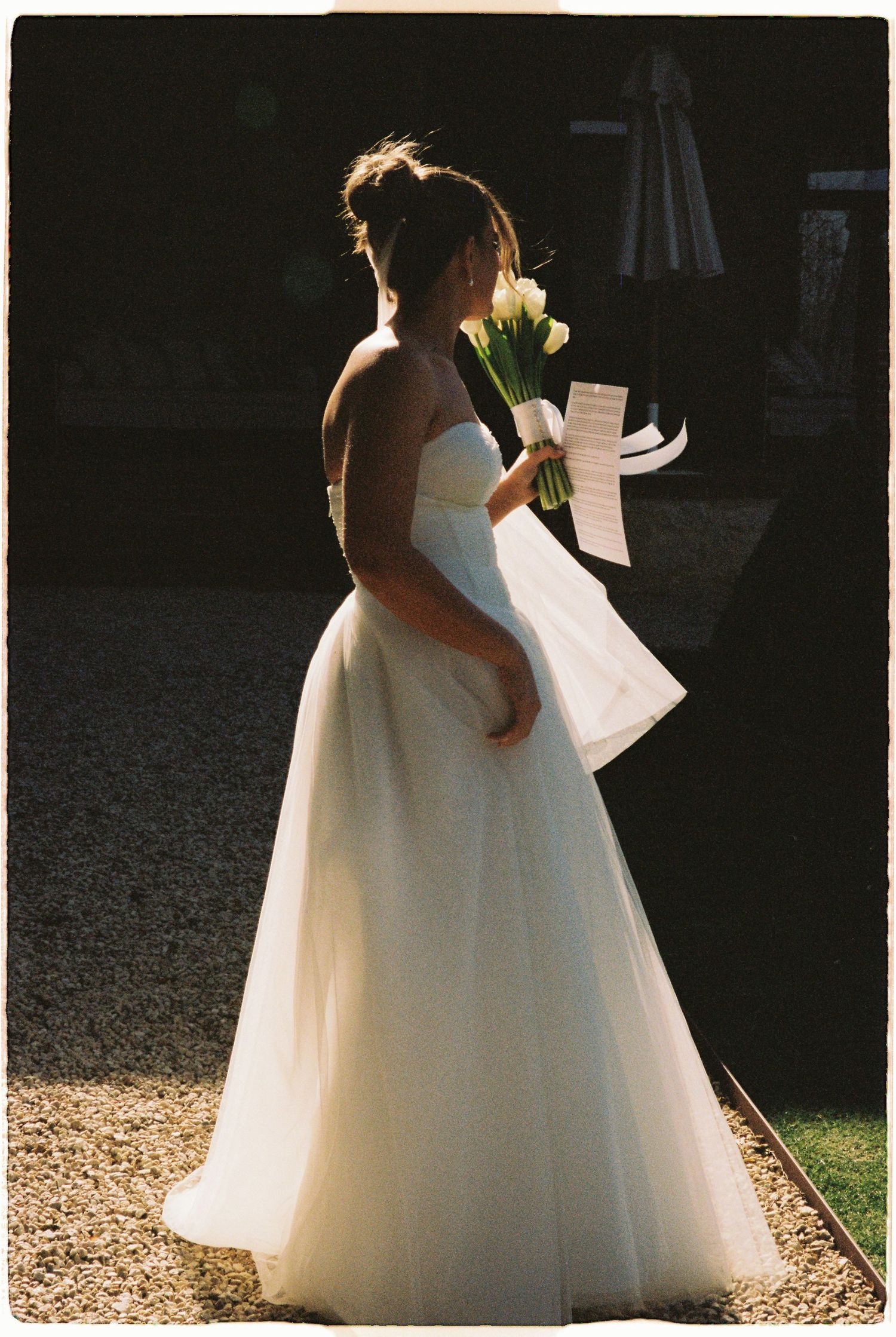 Bride in strapless white ballgown silhouetted in sunlight holding bouquet and papers.