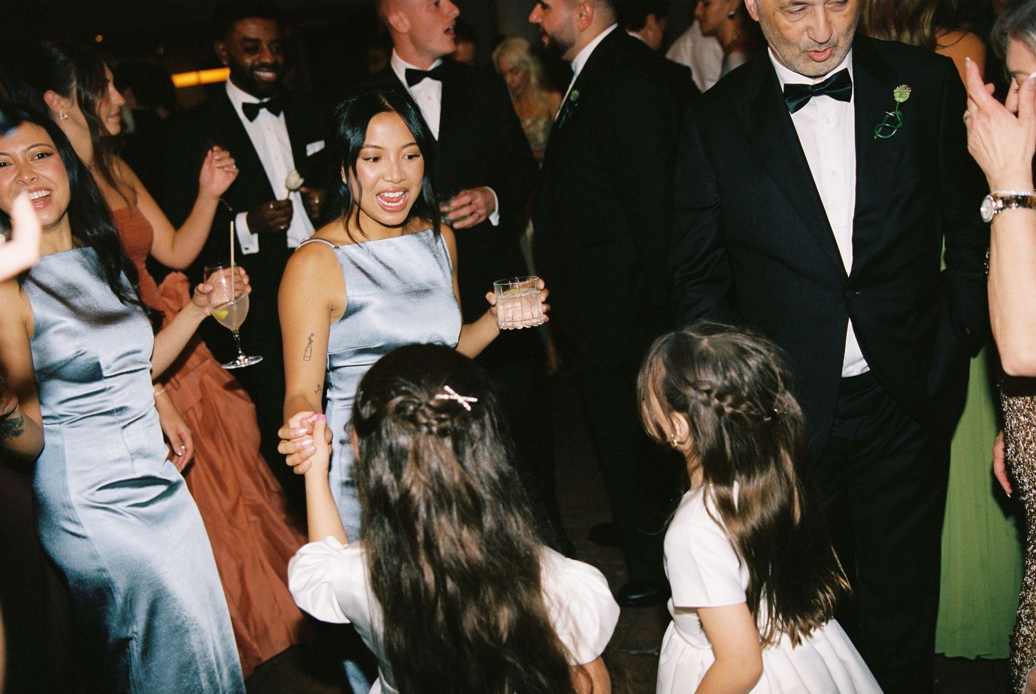 Bridesmaid in blue satin dress dancing with flower girls at wedding reception.