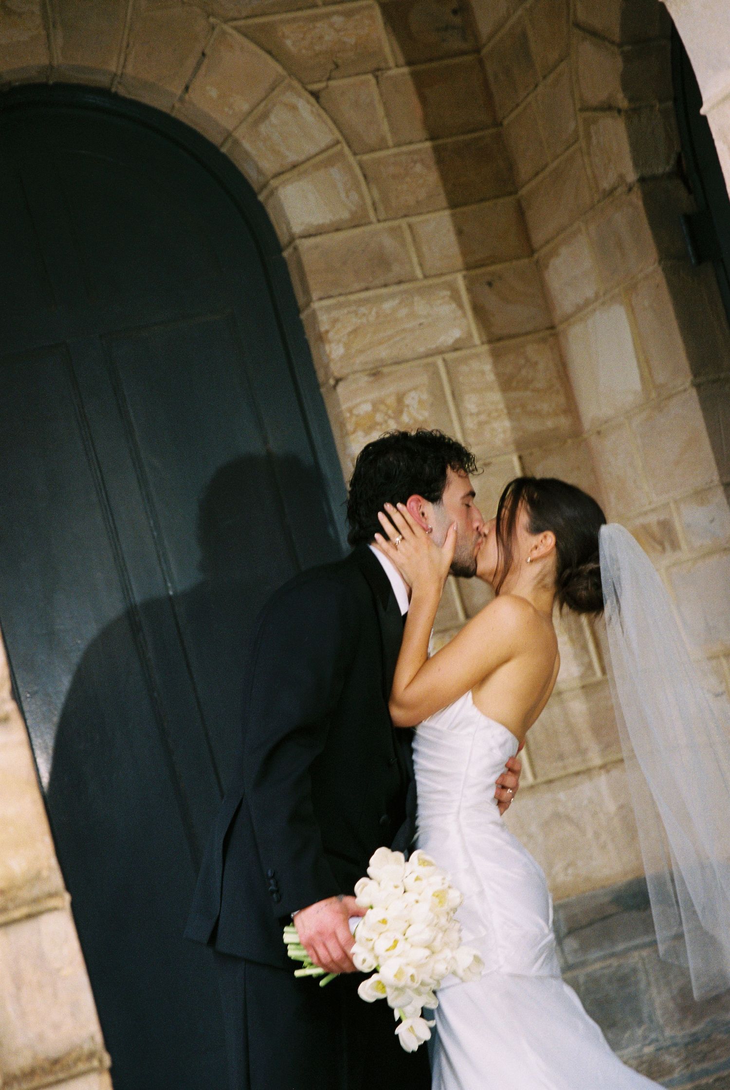 Bride and groom kiss passionately outside stone church doorway, bride holding white floral bouquet.