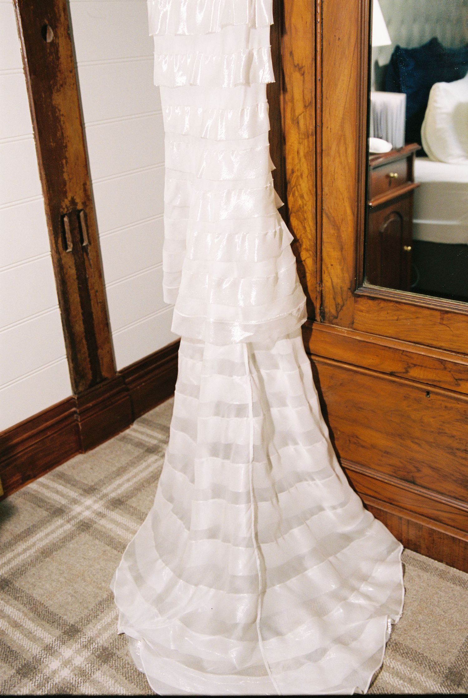 Close-up of a white ruffled tiered wedding dress hanging near a wooden wardrobe door.