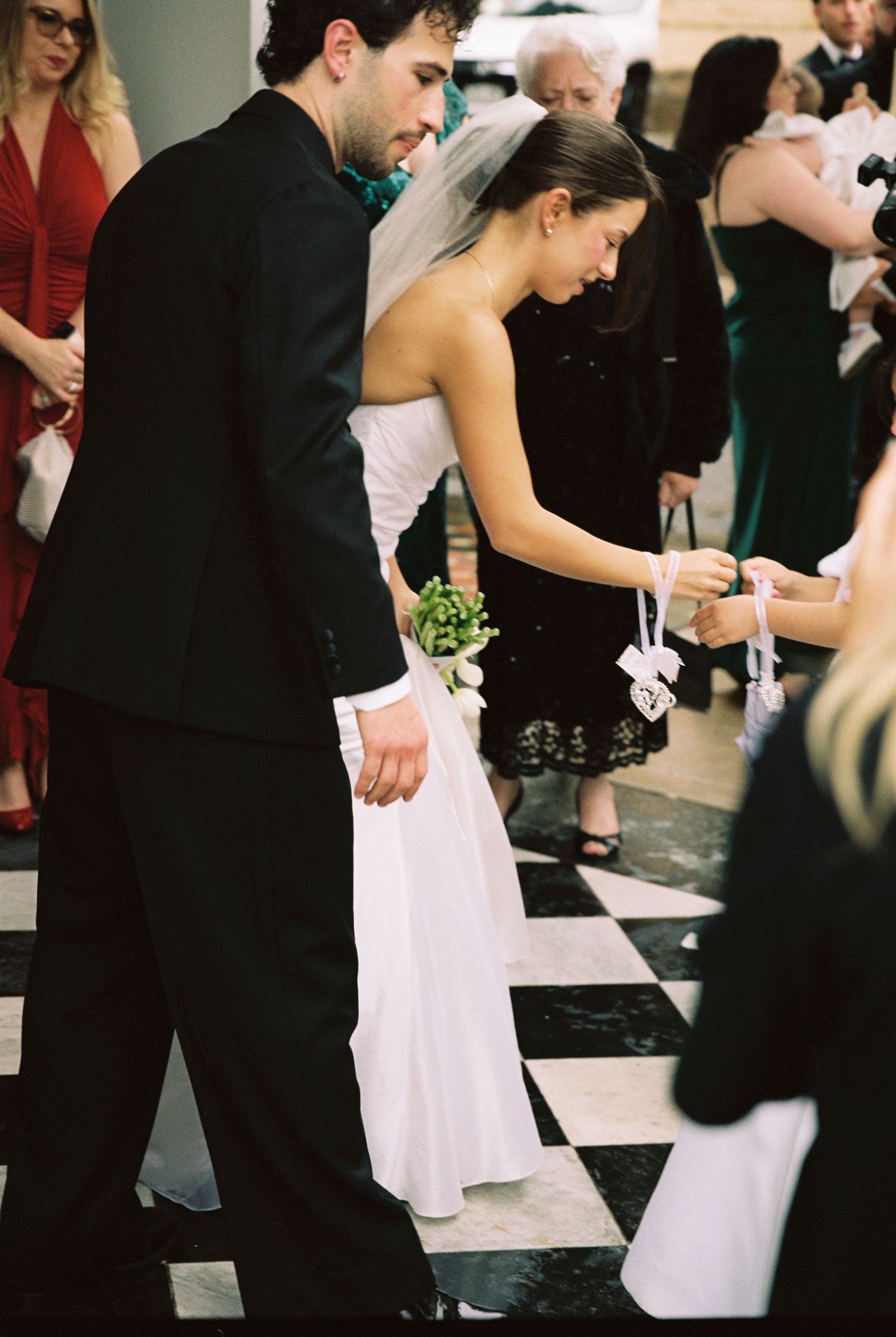 A bride in a white gown and groom in a black suit walk down the aisle on a checkered floor at their wedding.