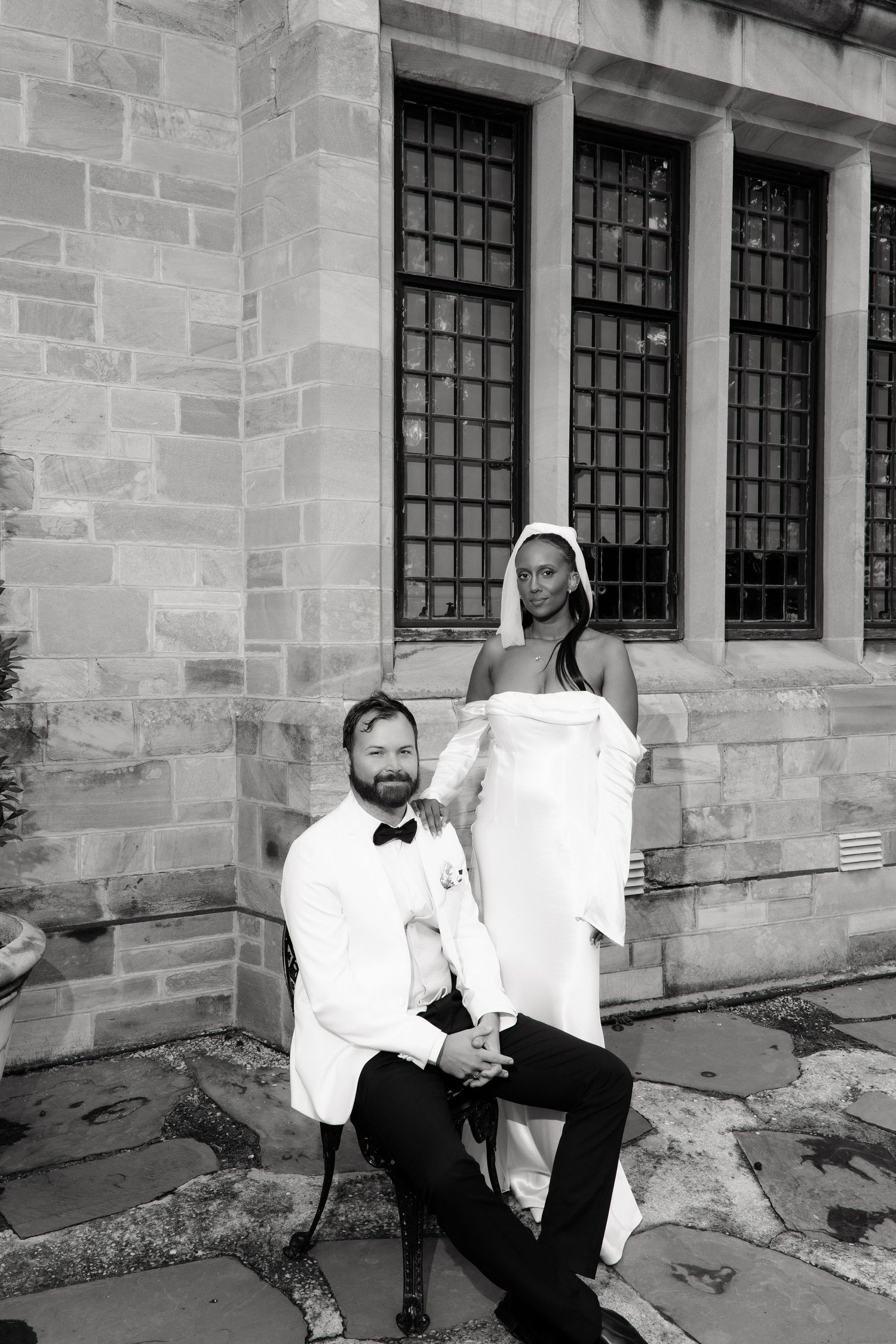 Black and white wedding photo of groom in white tuxedo seated before bride in strapless gown by stone building.