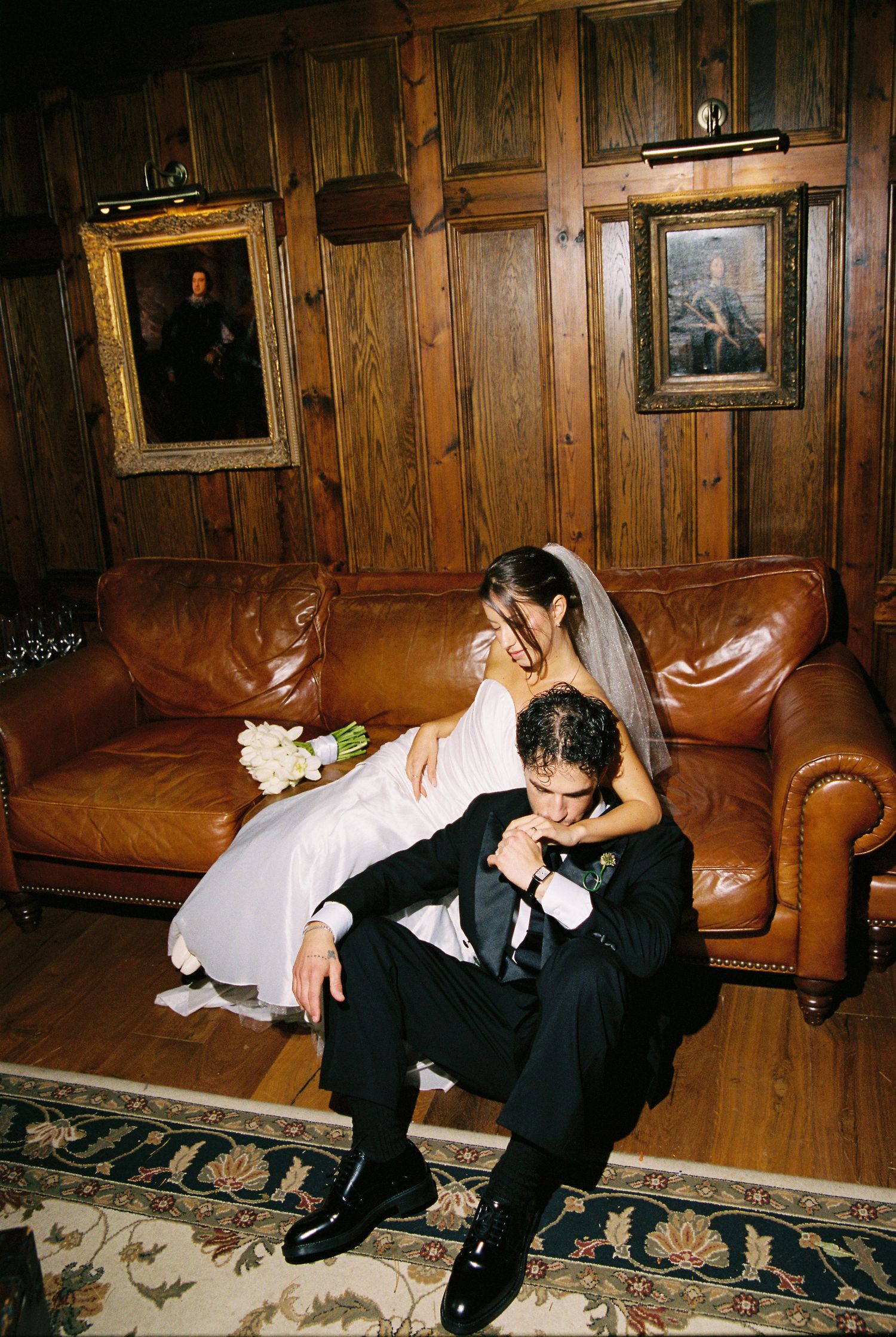 Bride and groom share intimate moment on leather sofa, surrounded by wood-paneled walls and classic portrait paintings.