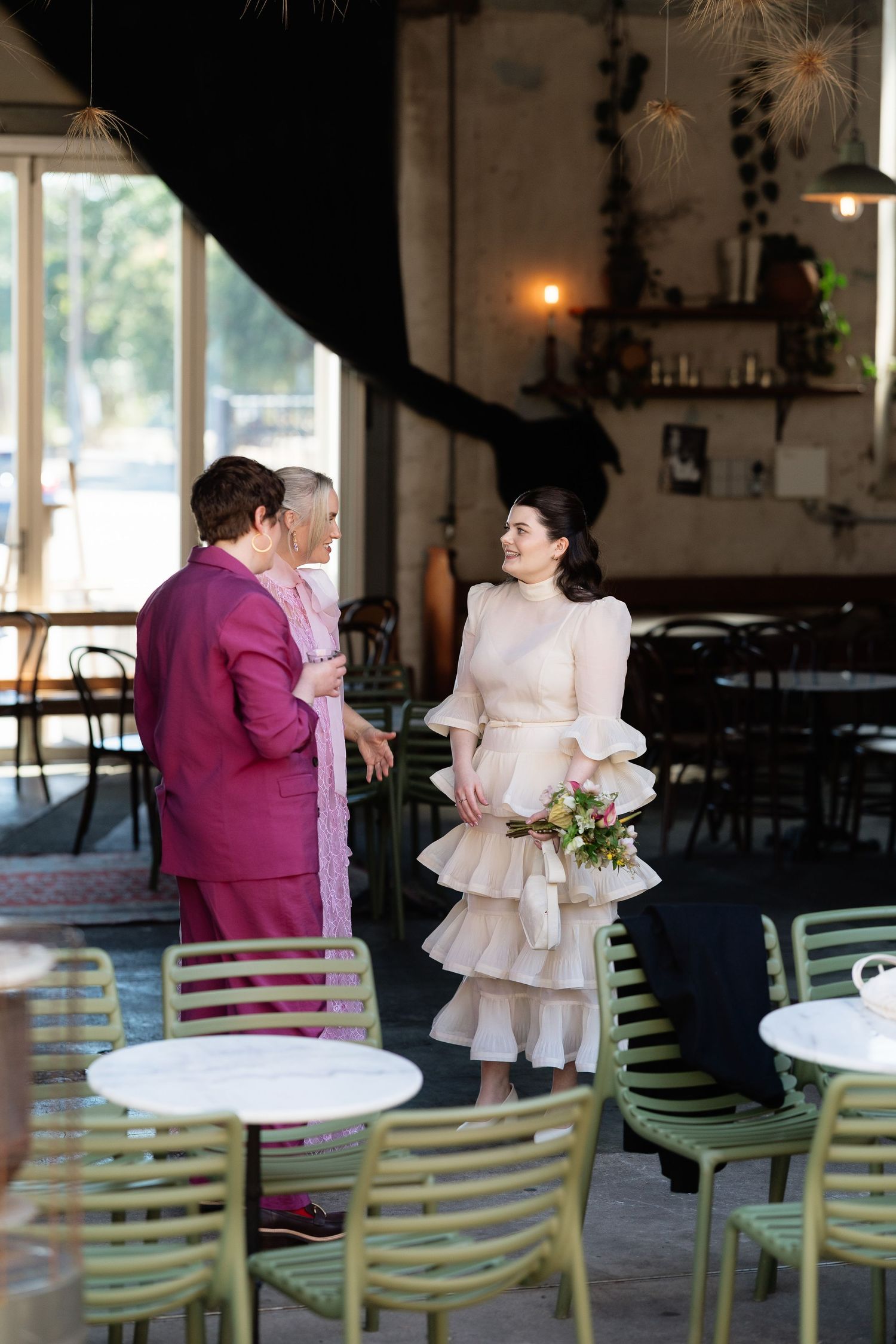 Bride in tiered ruffle dress holding bouquet smiles at guest in pink suit inside elegant restaurant venue.
