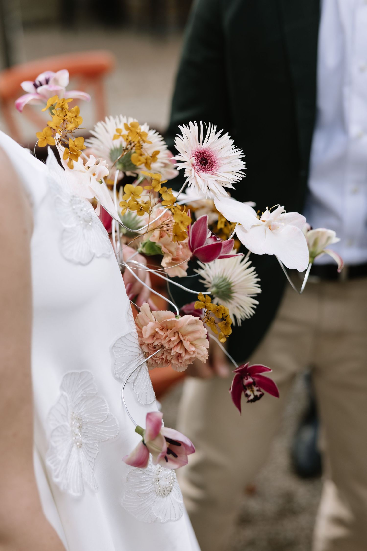 Bride holding a wild wedding bouquet with pink gerbera, yellow orchids, and burgundy blooms against white lace dress.