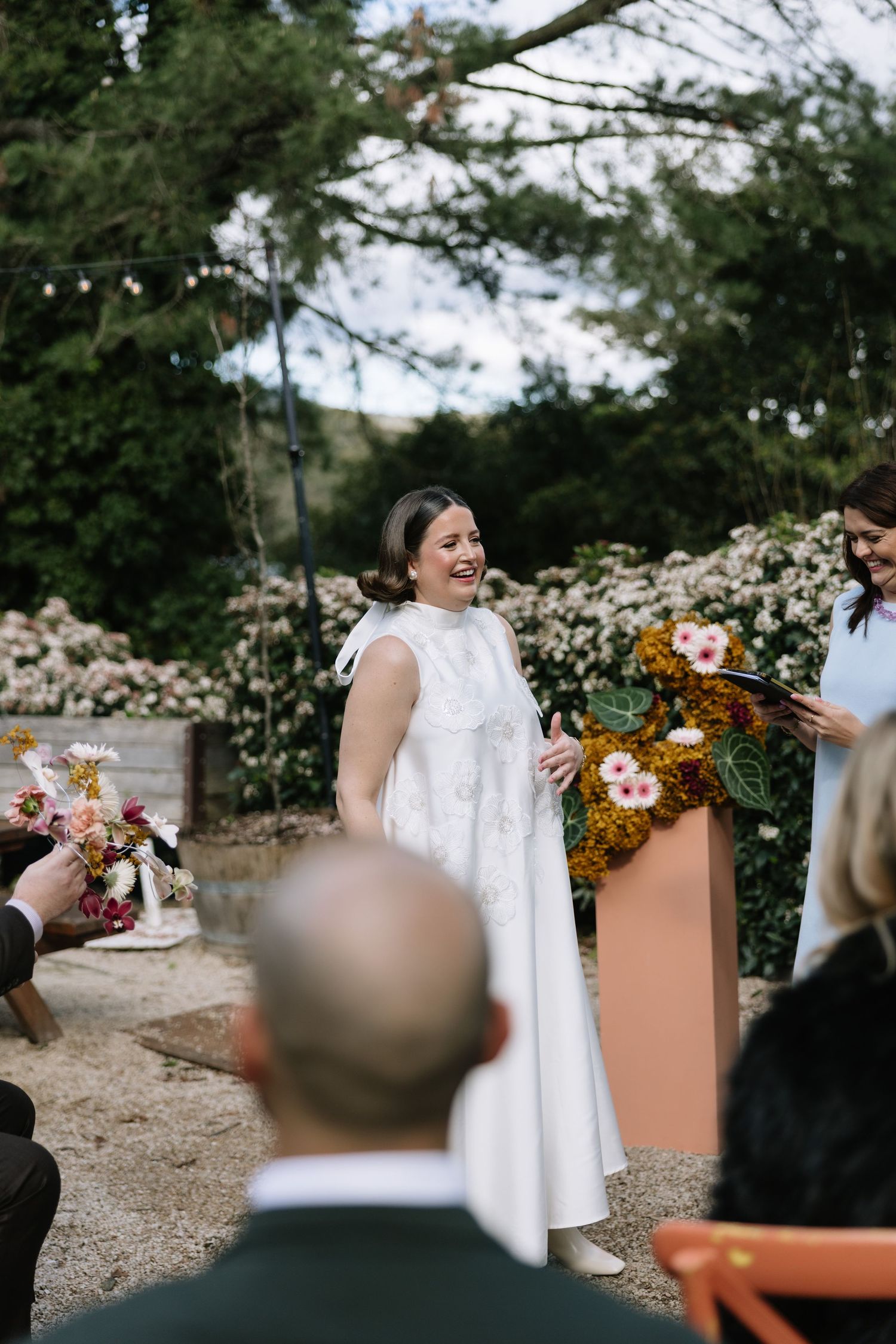 Bride in white dress laughing at outdoor wedding ceremony with floral arrangements and guests seated.