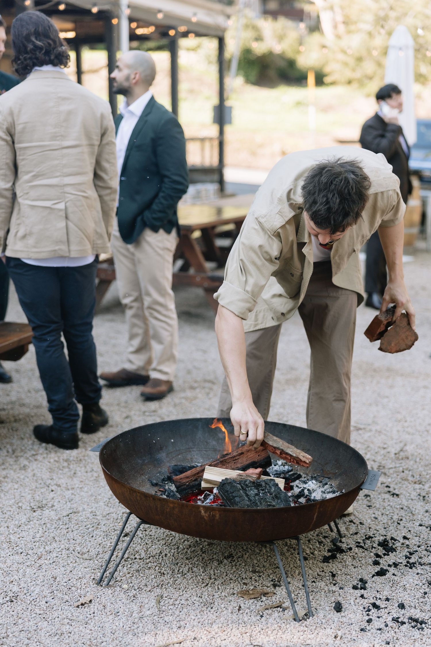 Man tending a fire pit at an outdoor gathering with guests in smart casual attire standing in the background.