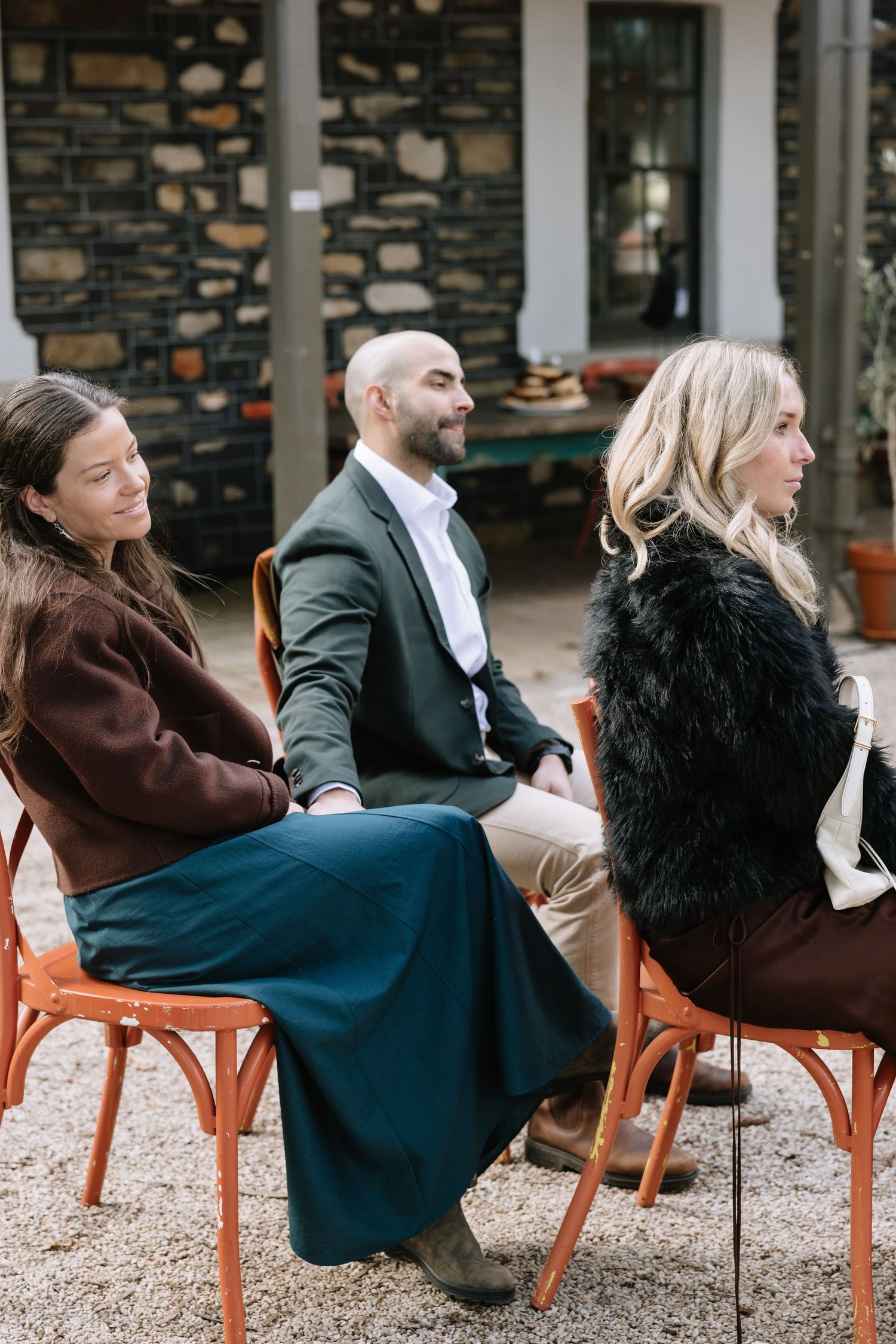 Three guests seated outdoors on orange chairs at a wedding ceremony, wearing elegant attire against a stone wall backdrop.