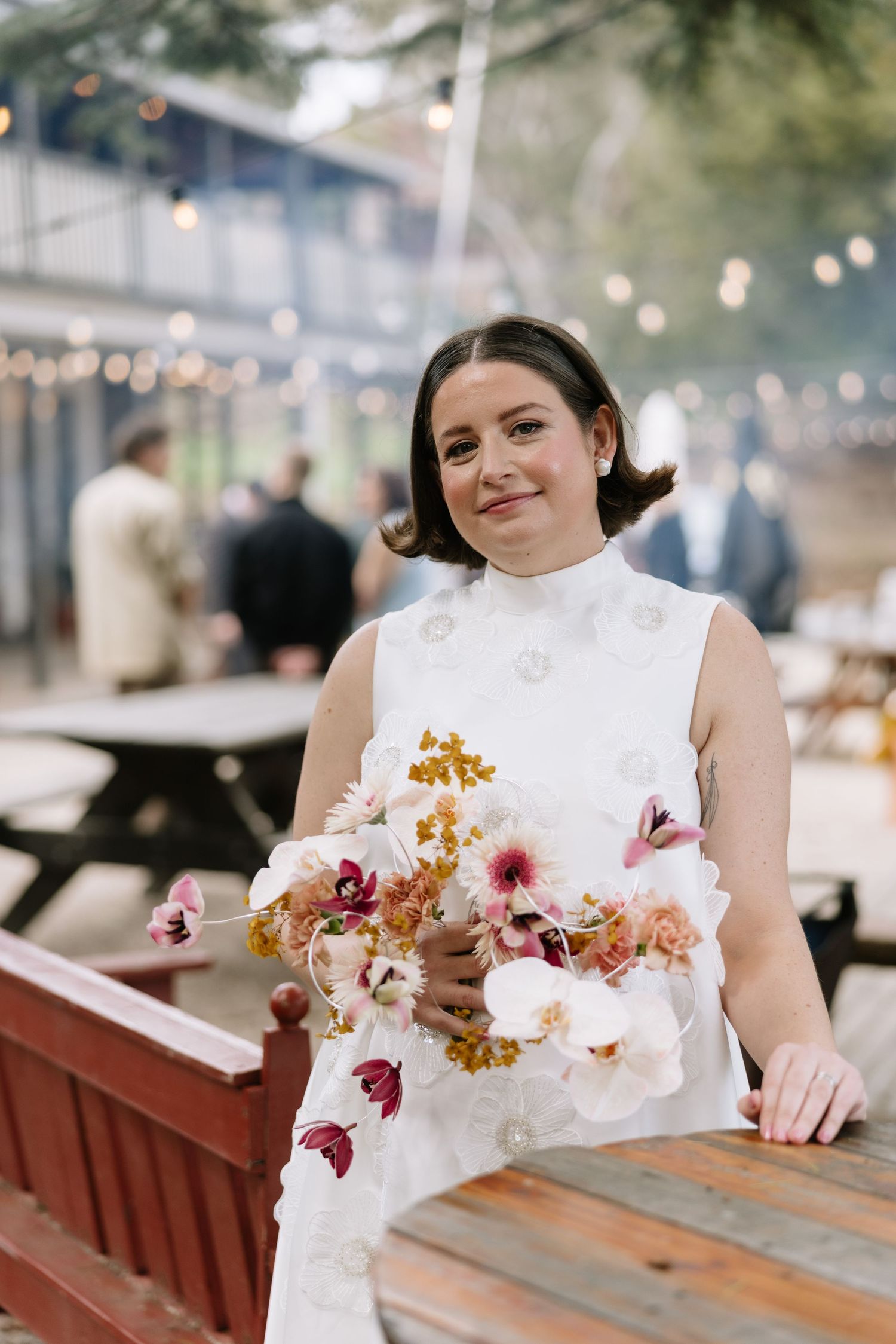 Bride in white floral dress holding a colorful bouquet of orchids and wildflowers at an outdoor wedding reception.