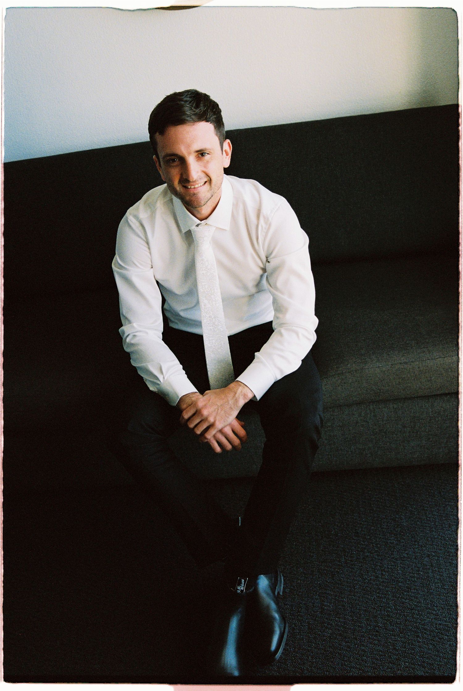 Smiling groom in white shirt and tie sitting relaxed on dark sofa before wedding ceremony.