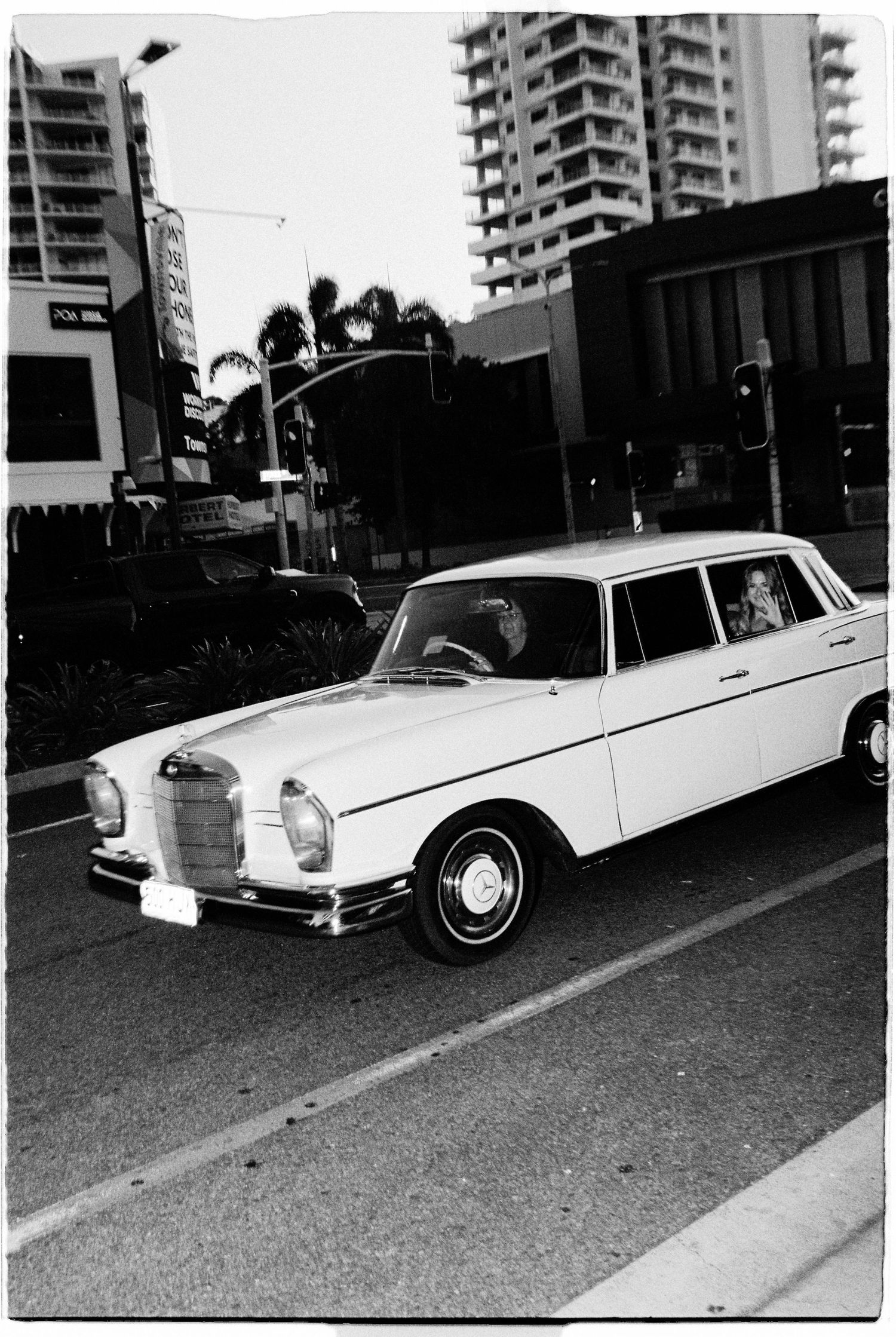 Black and white photo of vintage white Mercedes-Benz driving through urban intersection.