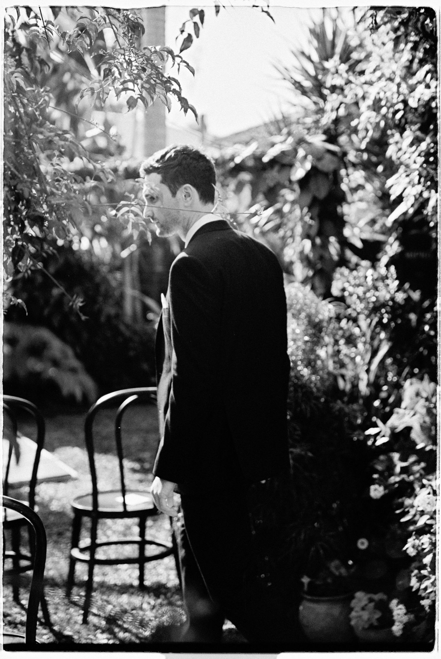 Black and white photo of a groom standing in a lush garden surrounded by chairs before the wedding ceremony.