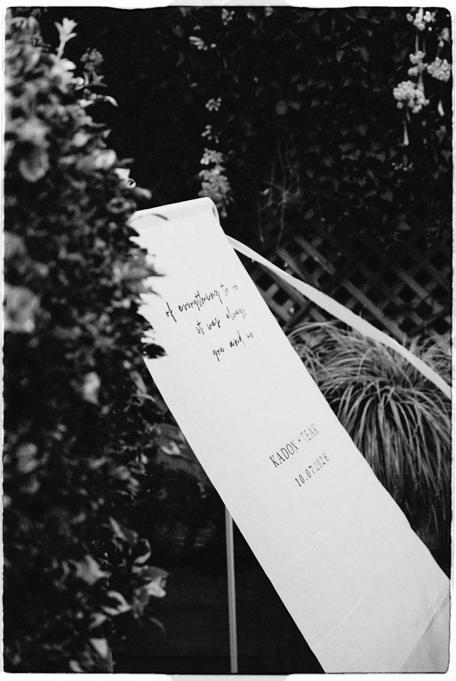 Black and white photo of wedding ceremony ribbon sign with handwritten vows among garden greenery.