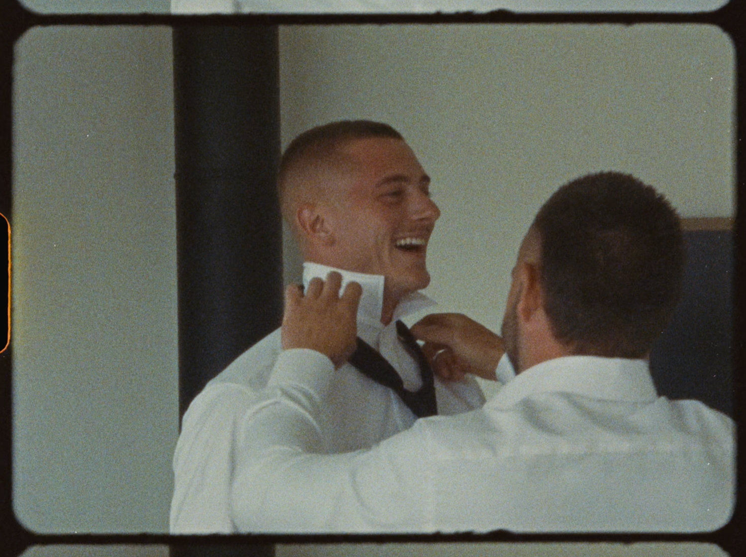 Groomsman laughing while another helps adjust his tie during wedding day preparations.