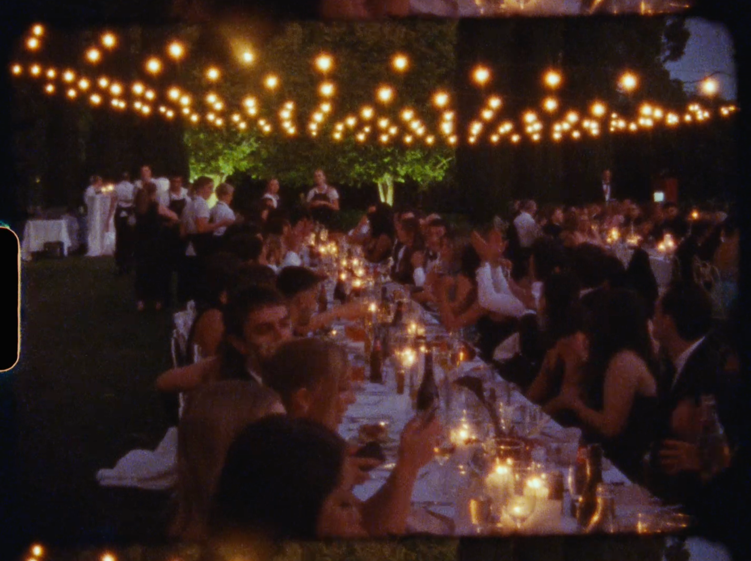 Romantic outdoor wedding reception with string lights and candlelit long dining tables filled with elegantly dressed guests.
