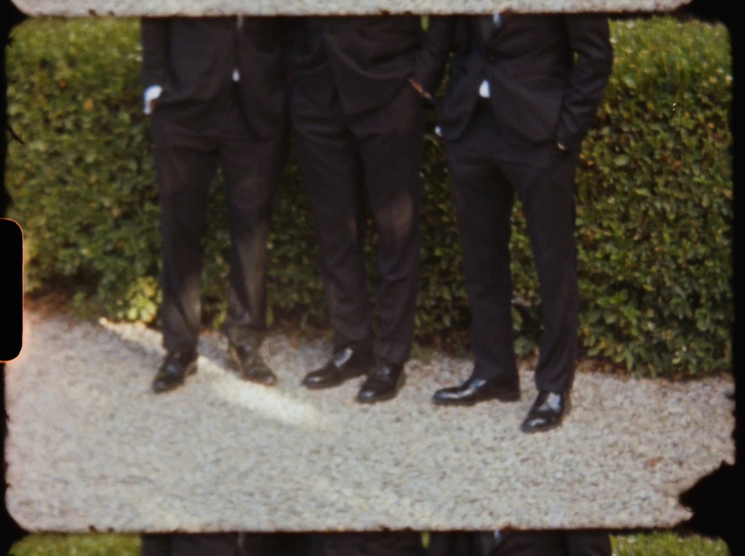 Vintage film-style photo of three groomsmen in black suits and dress shoes standing on gravel.