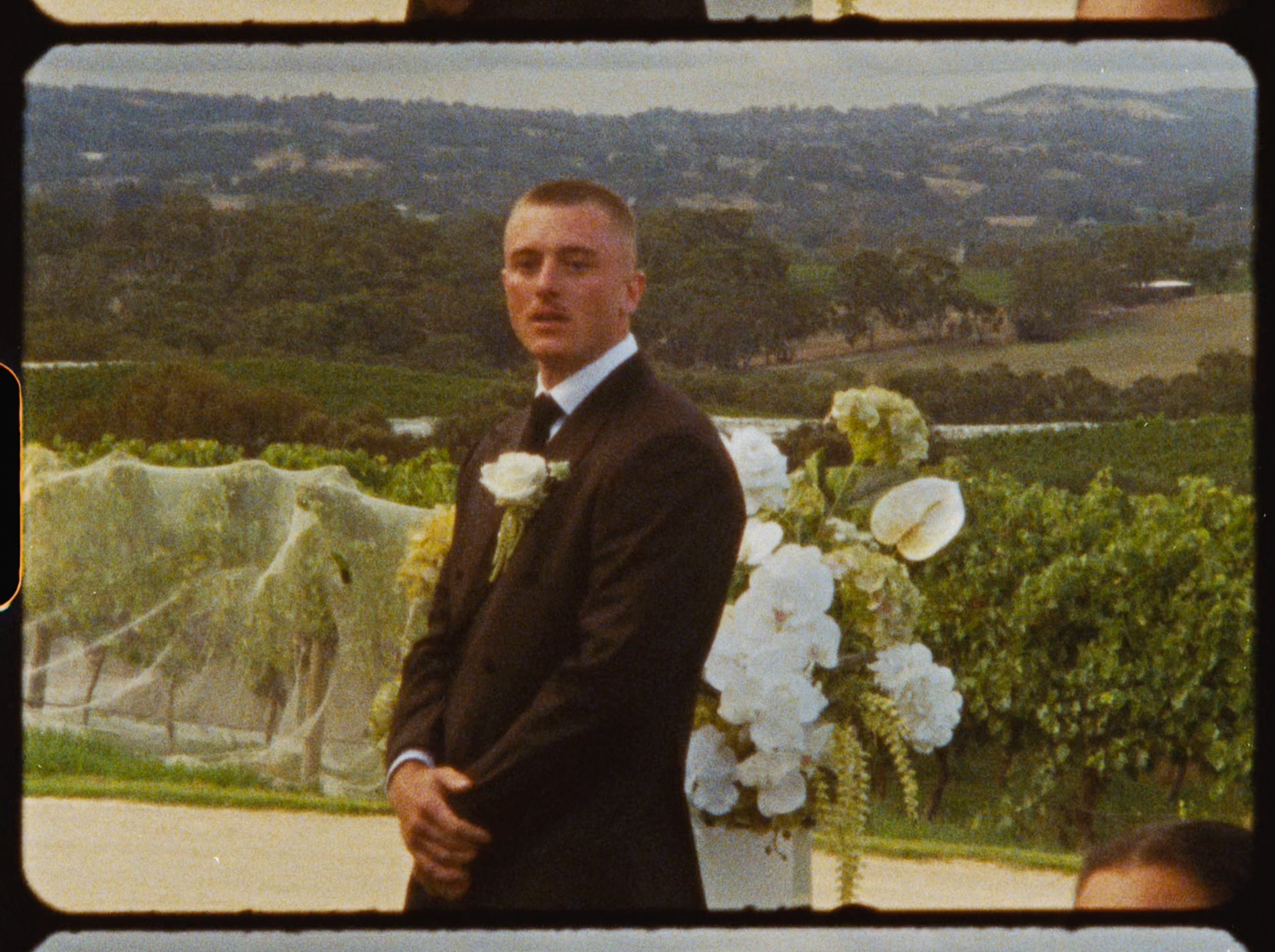 Groom in black tuxedo standing at outdoor vineyard wedding altar with white floral arrangements.