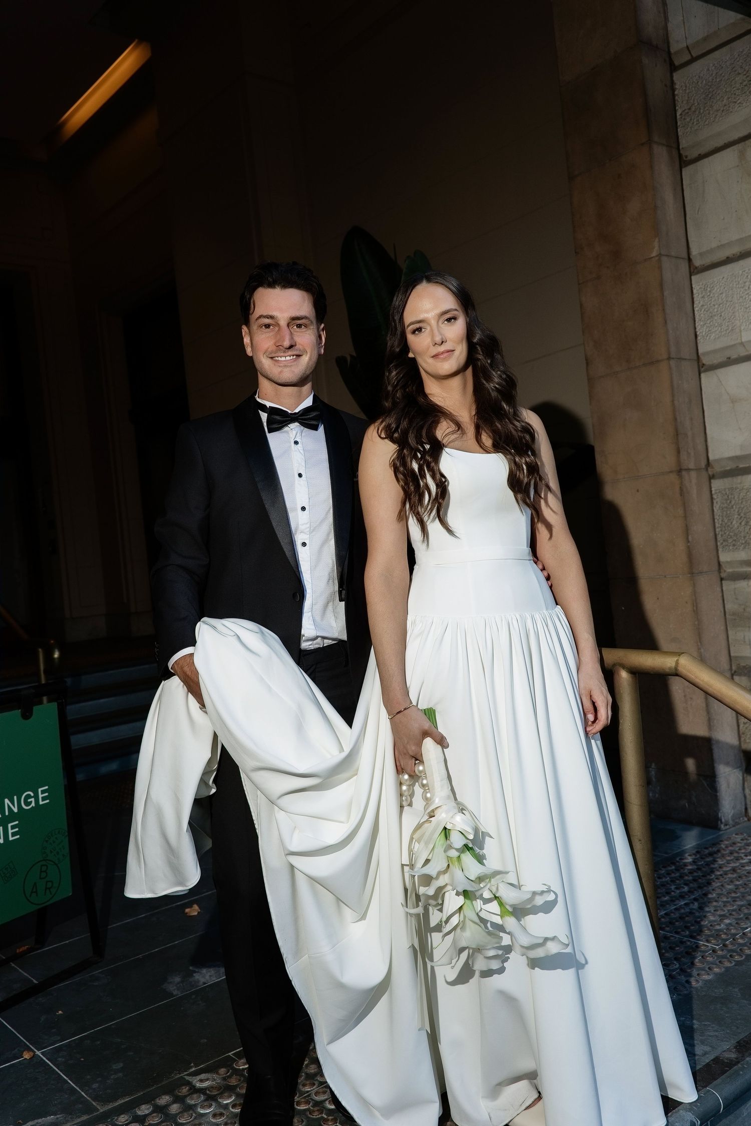 Smiling bride and groom posing outside a grand building entrance, groom holding bridal train.