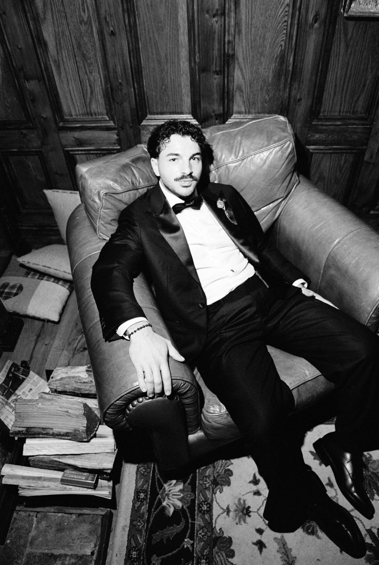 Stylish groom in black tuxedo relaxing on leather sofa in black and white portrait photo.