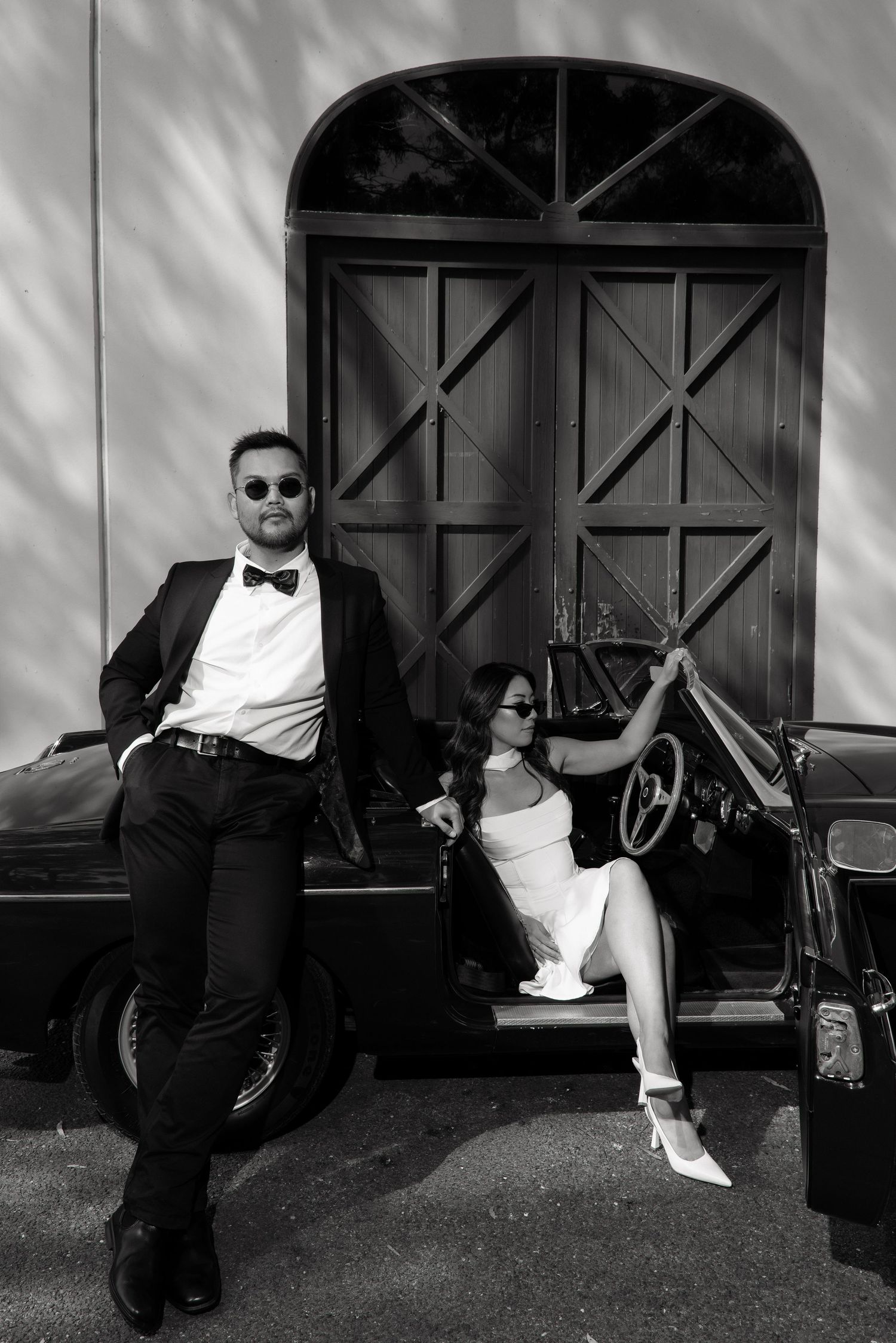 Stylish couple in black and white, man in tuxedo and woman in white dress by vintage car.