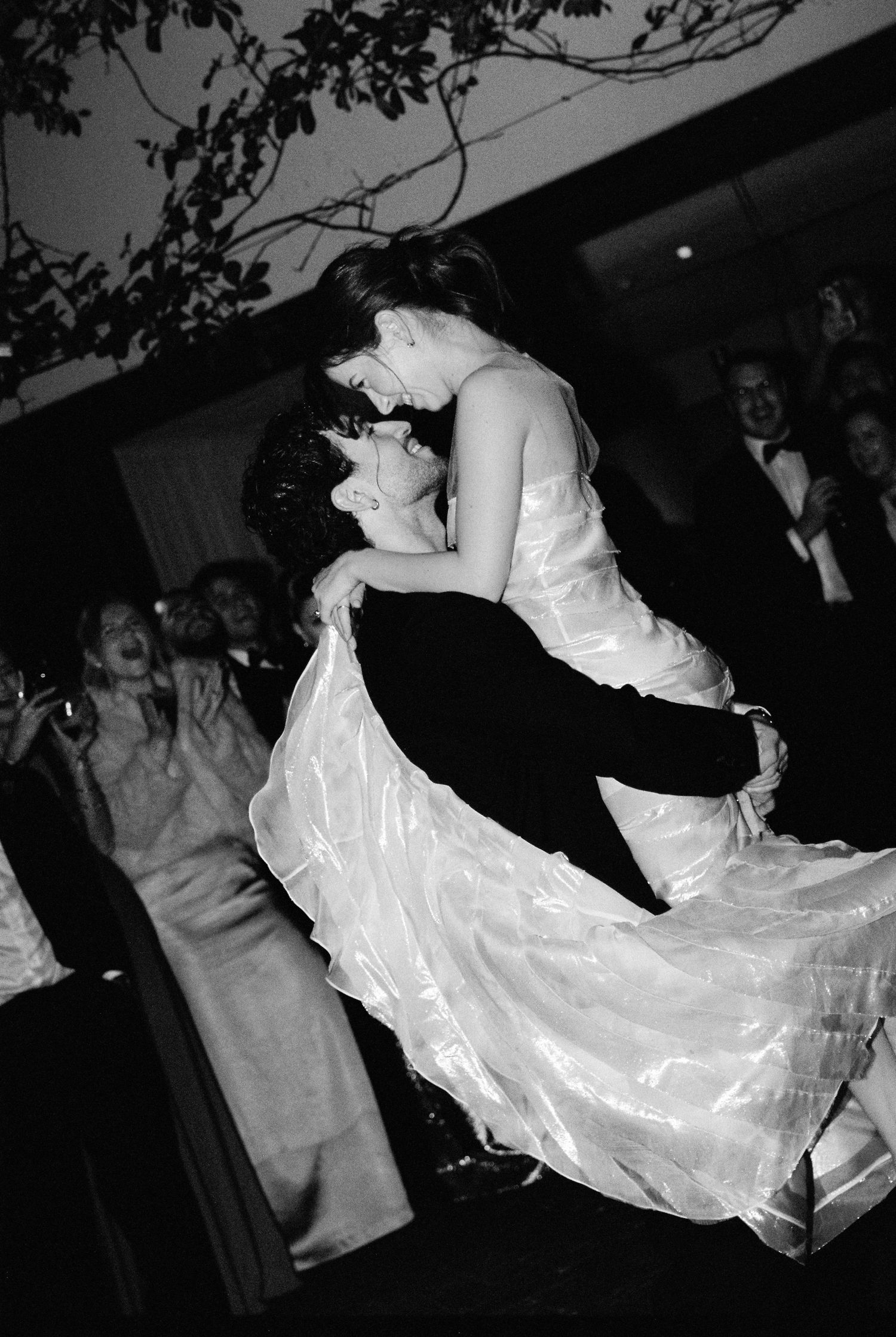 Groom lifts bride in wedding dress during energetic first dance while guests cheer in background.