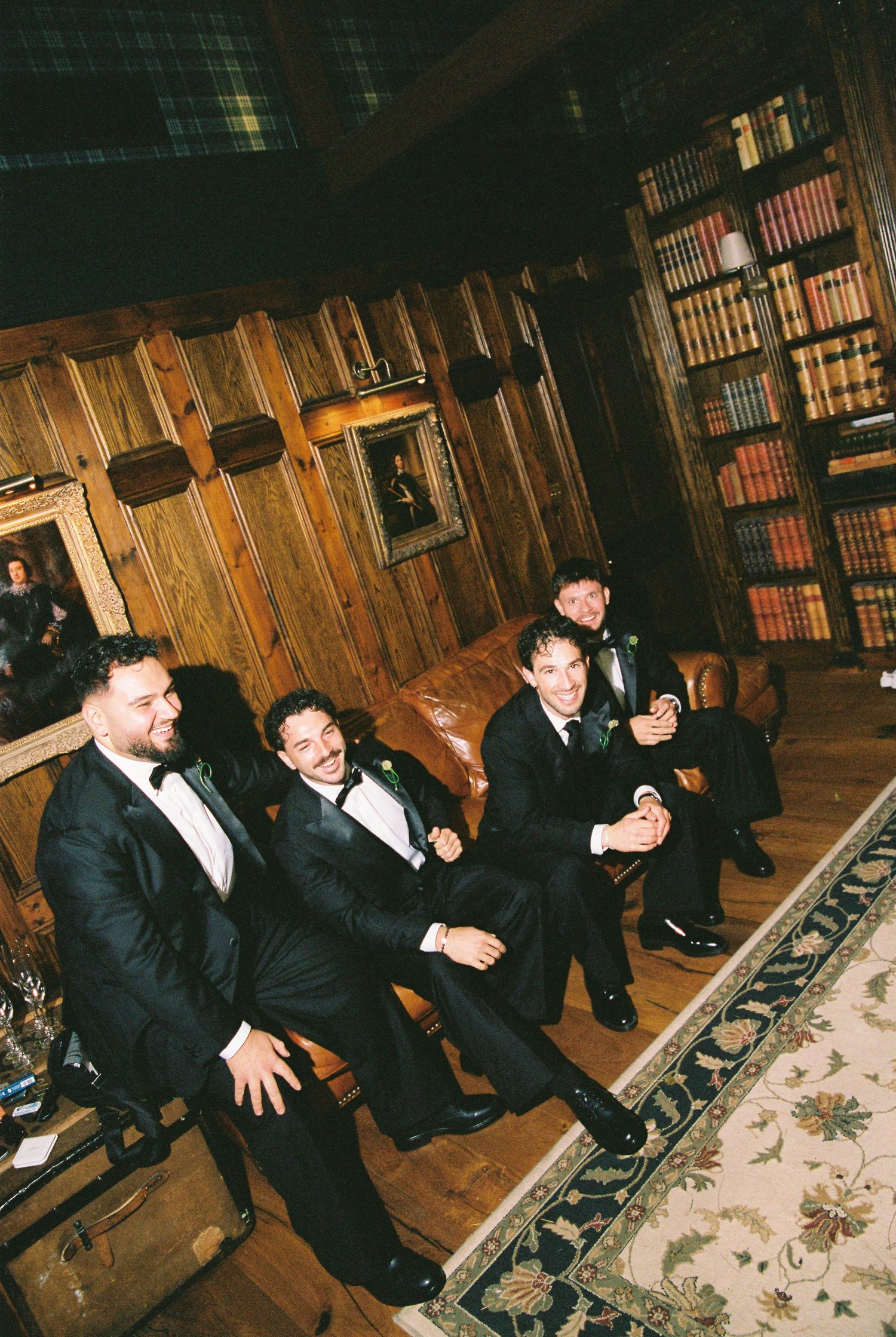 Four groomsmen in black tuxedos laughing together in a wood-paneled library.
