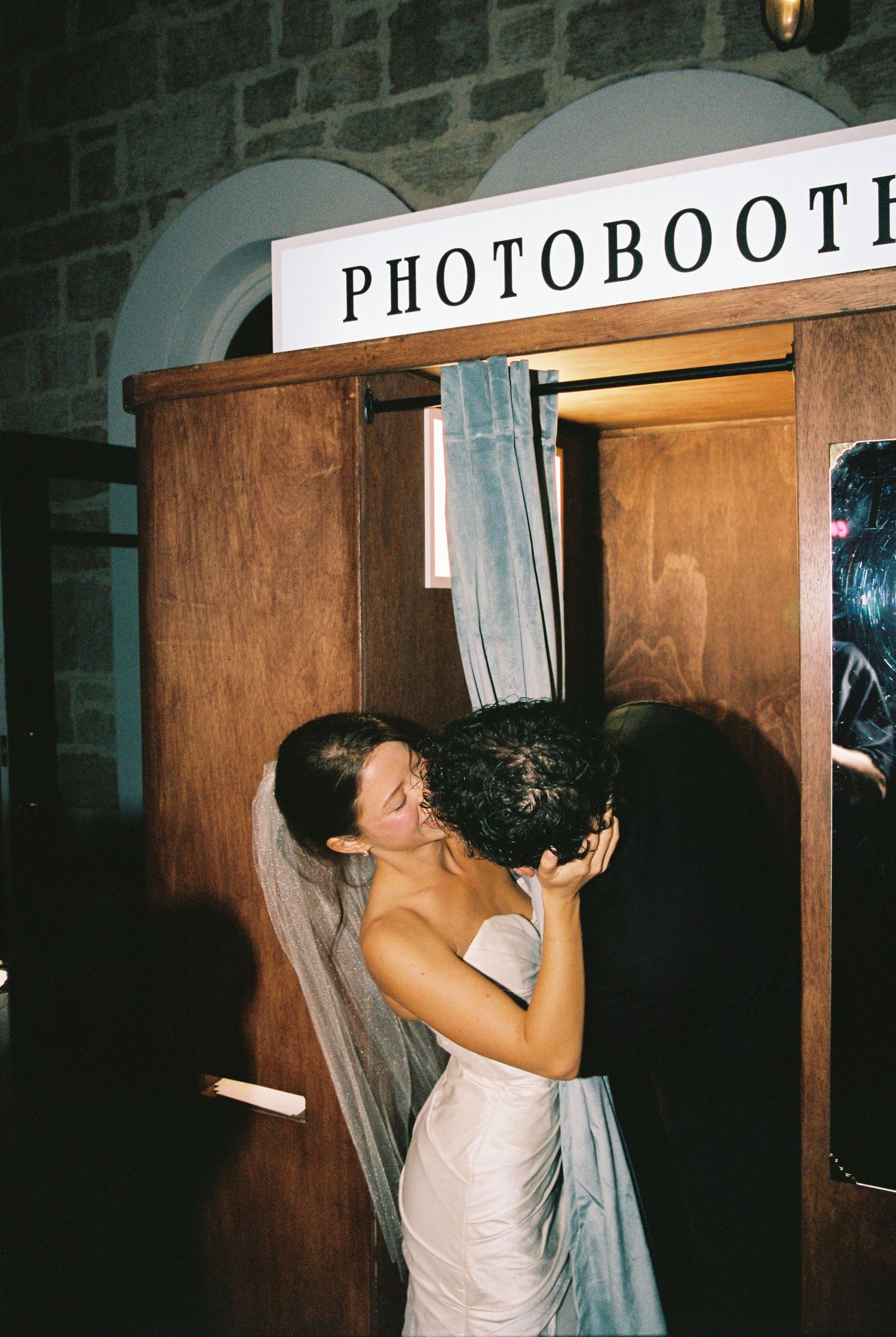 Bride kissing partner in a vintage wooden photo booth at a wedding reception.