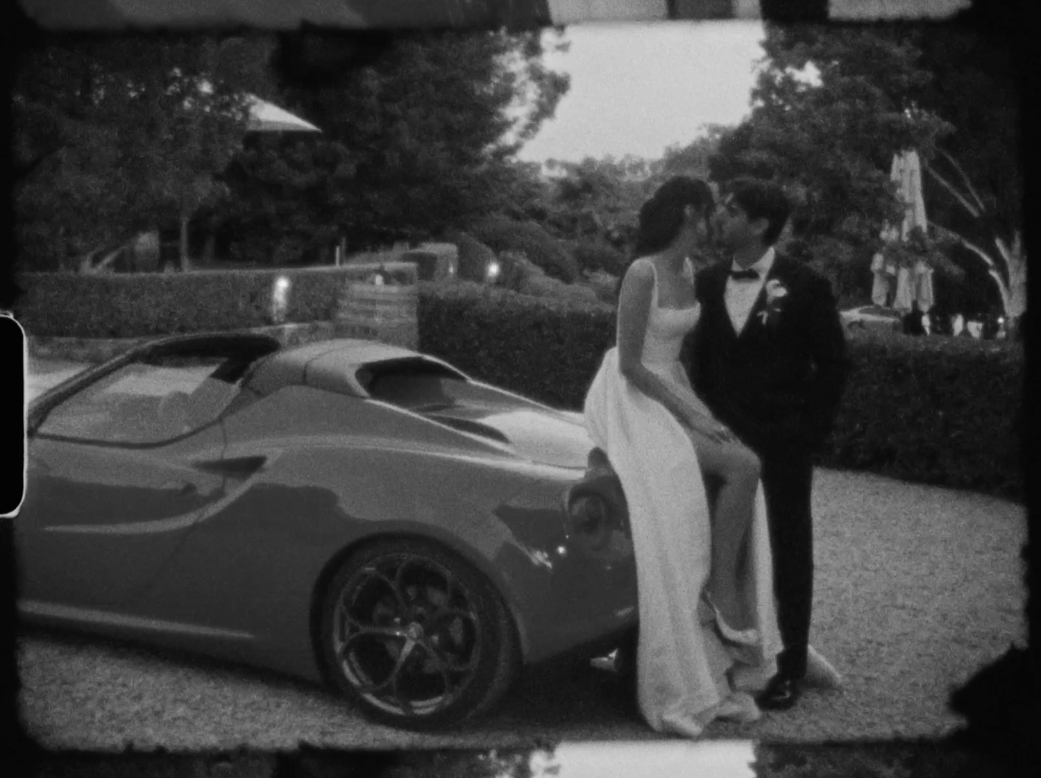 Bride and groom kissing beside a sleek sports car at their outdoor wedding venue in black and white.