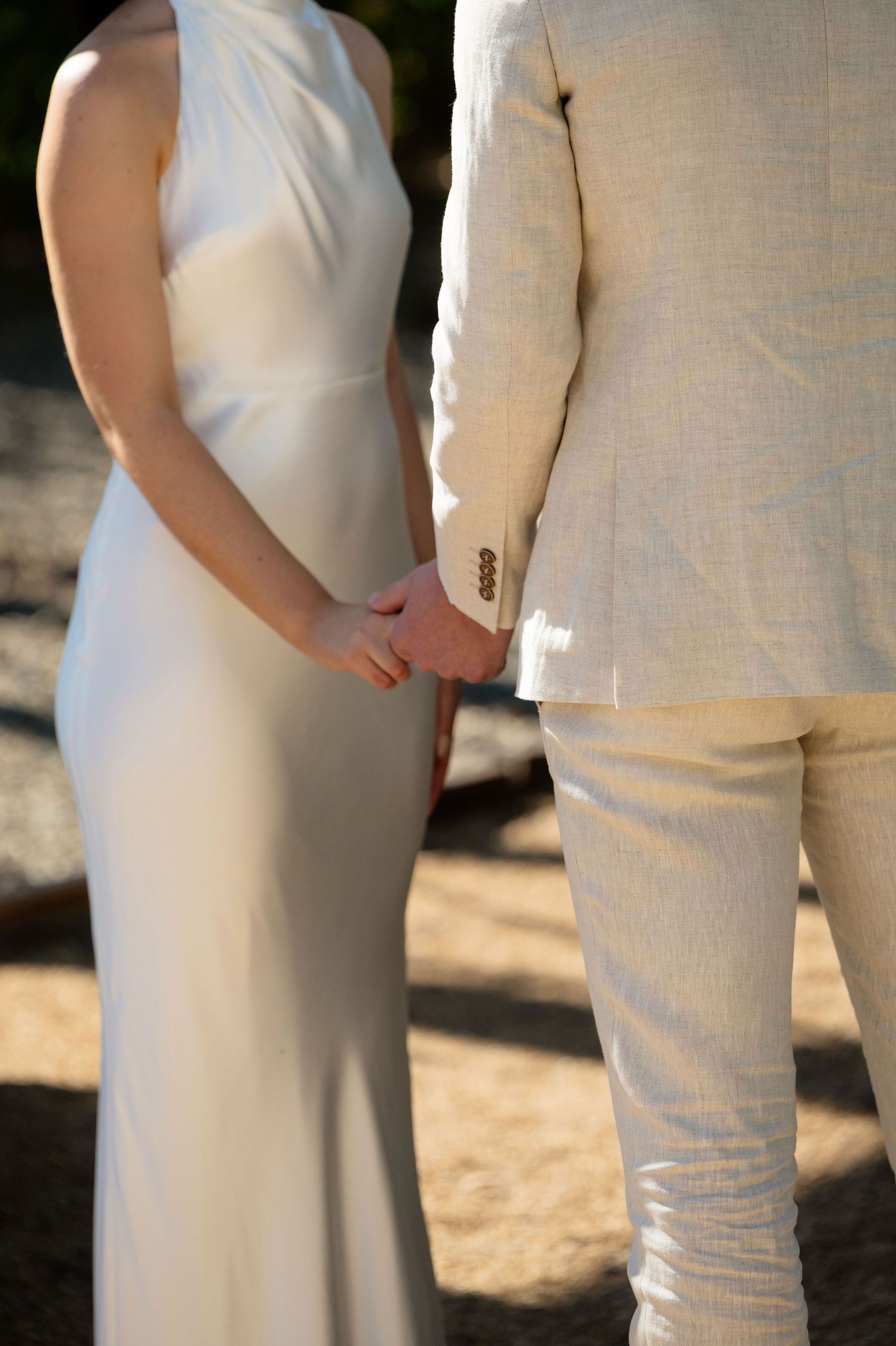 Bride in white sleeveless gown and groom in cream linen suit hold hands during outdoor wedding ceremony.