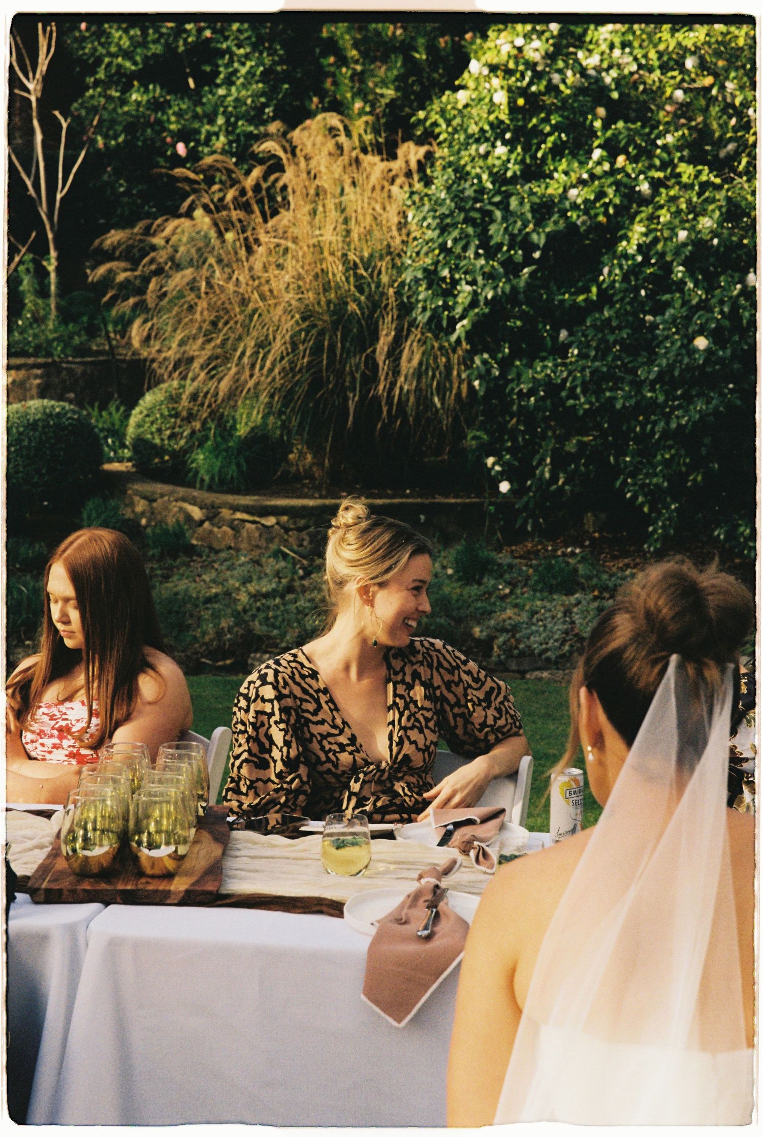 Bride in veil sits at outdoor garden wedding reception table with smiling guests in lush green surroundings.