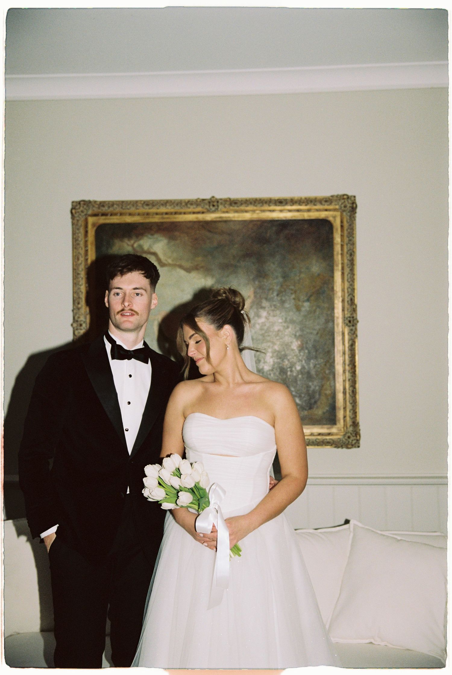 Bride and groom posing indoors before classic oil painting, groom in tuxedo, bride holding tulips.
