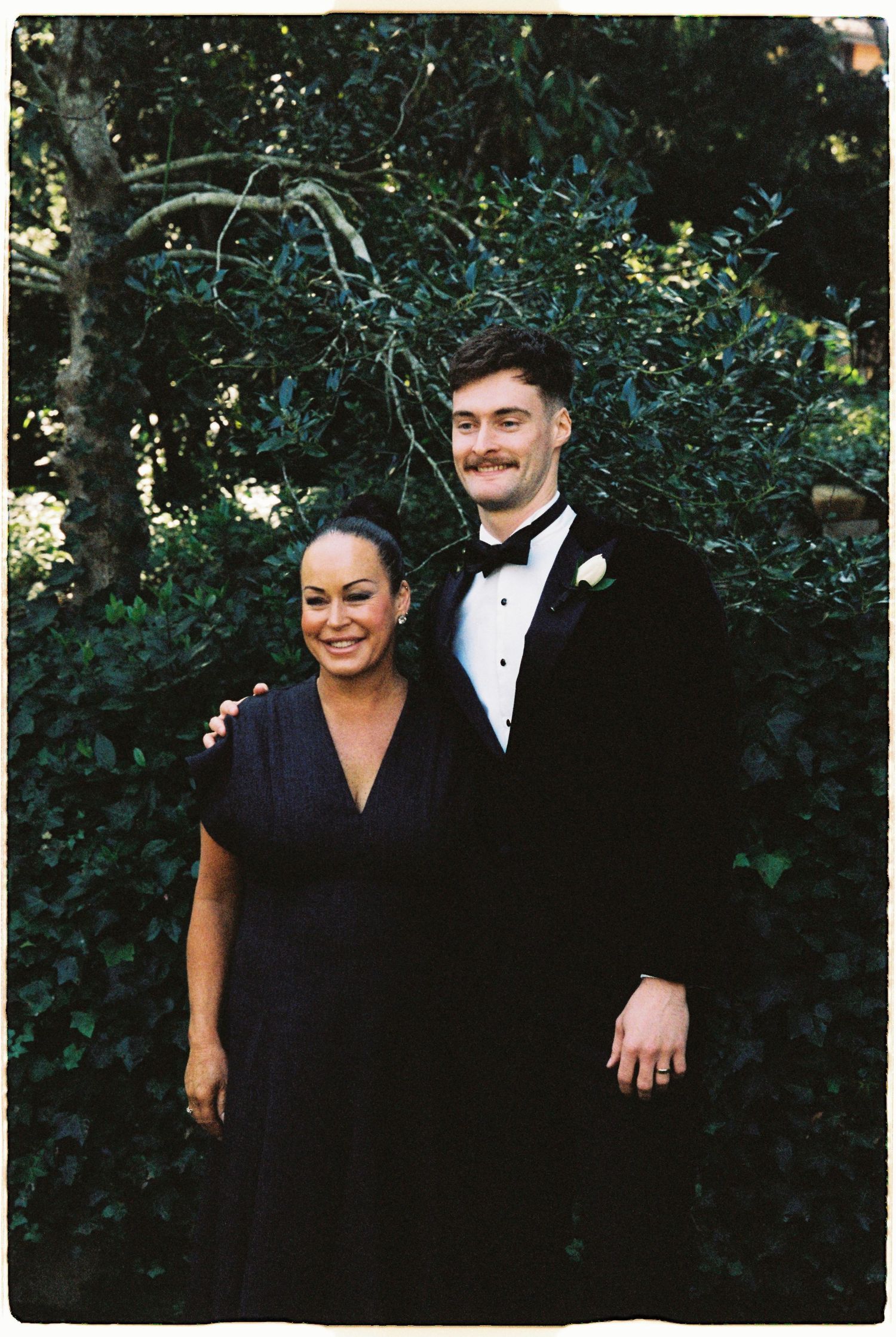 Young man in black tuxedo poses with a woman in a navy dress in front of lush green foliage.