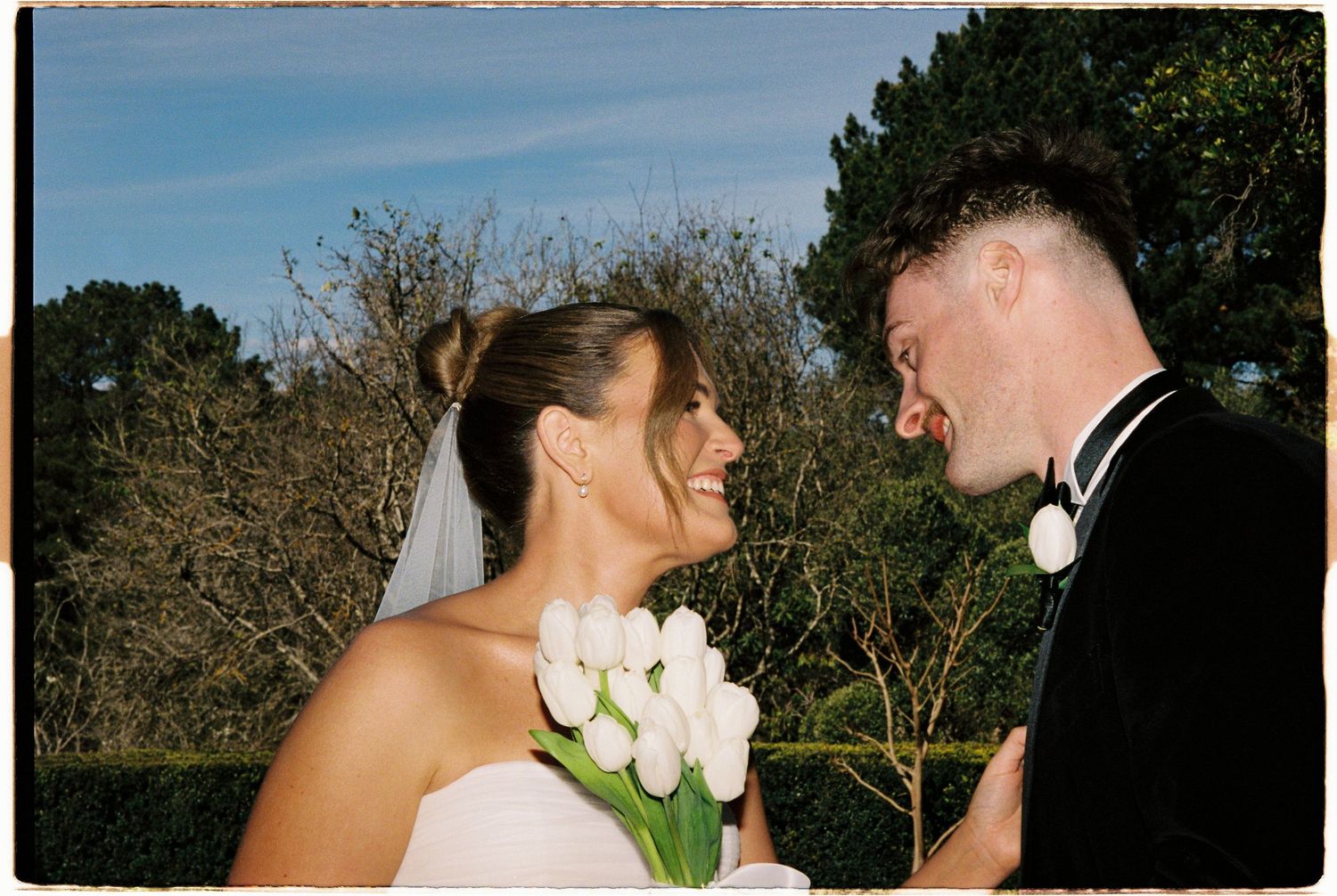 Smiling bride holding white tulip bouquet laughing with groom in black tuxedo at outdoor wedding.