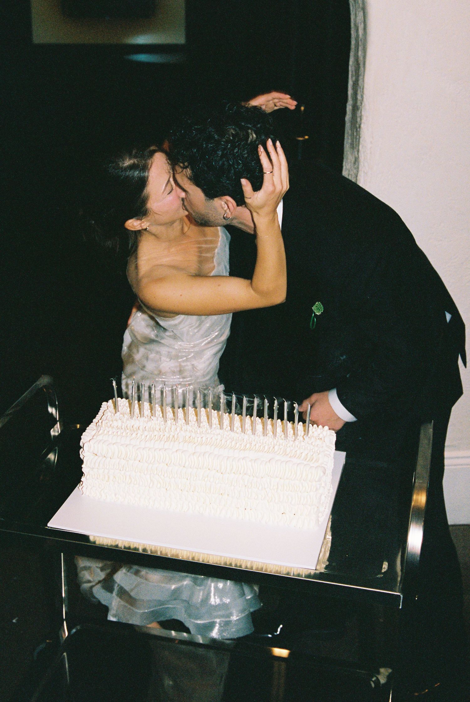 Newlyweds sharing a romantic kiss over their white wedding cake with candles.