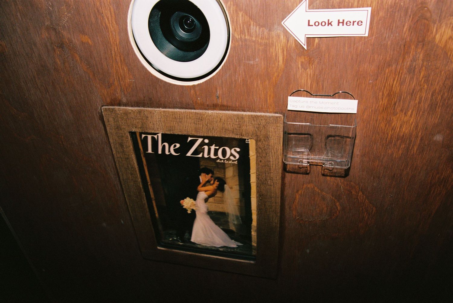 A vintage photo booth interior with a camera, arrow sign reading Look Here, and The Zitos wedding photo card on wood paneling.