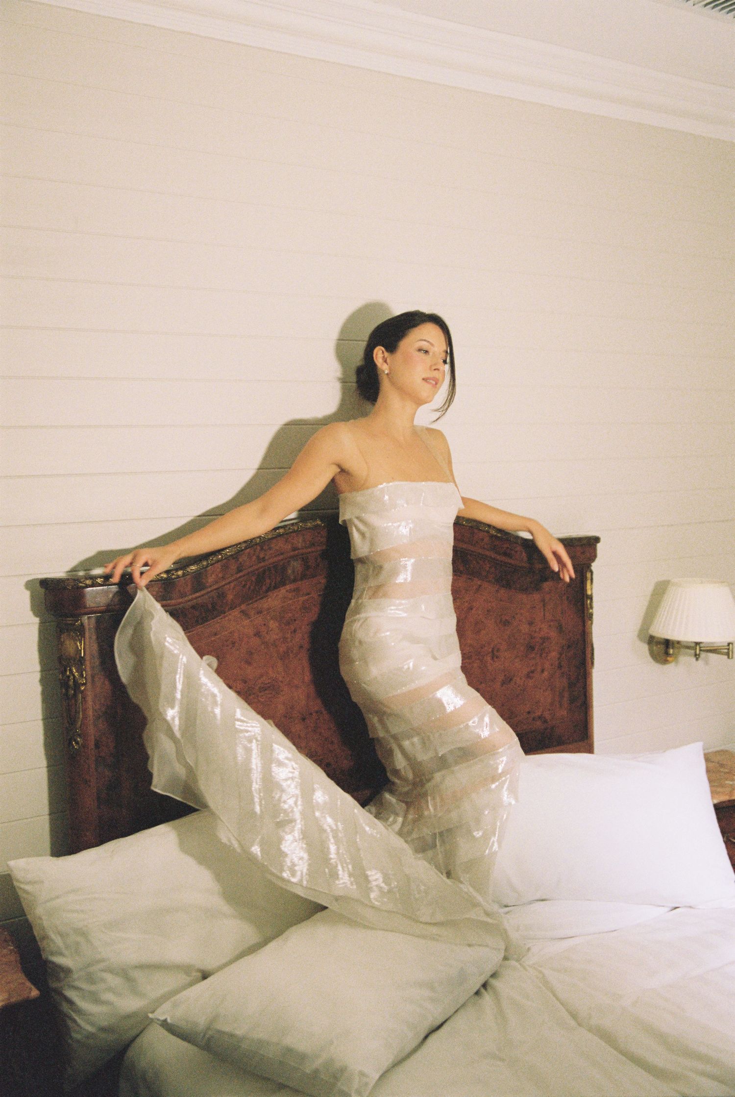 Bride in a flowing white strapless gown posing gracefully on a bed with an ornate wooden headboard.