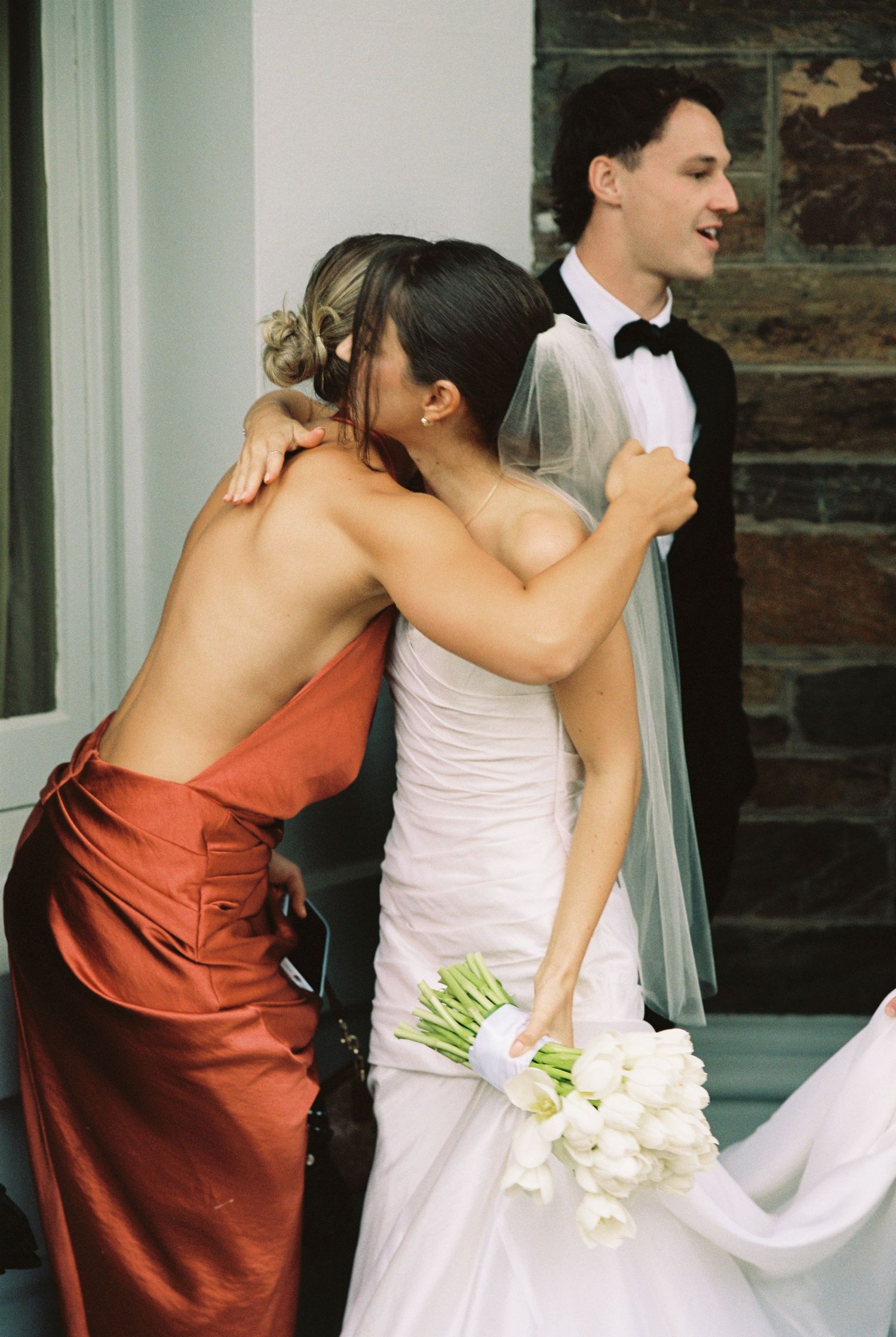 Bridesmaid in rust satin gown embraces bride in white wedding dress holding white tulip bouquet.