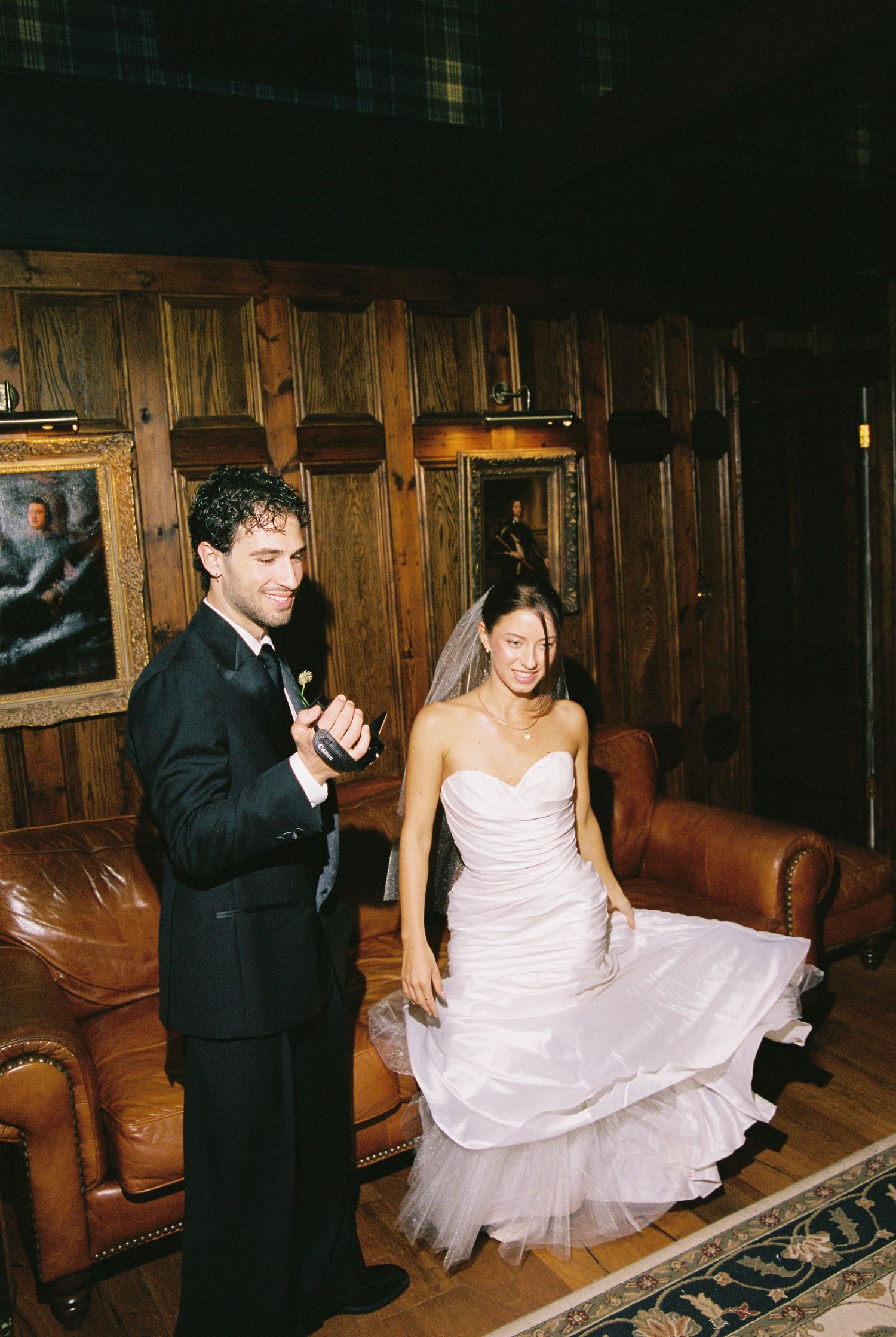 Bride in white strapless gown and veil laughs with groom in black tuxedo in wood-paneled wedding venue.