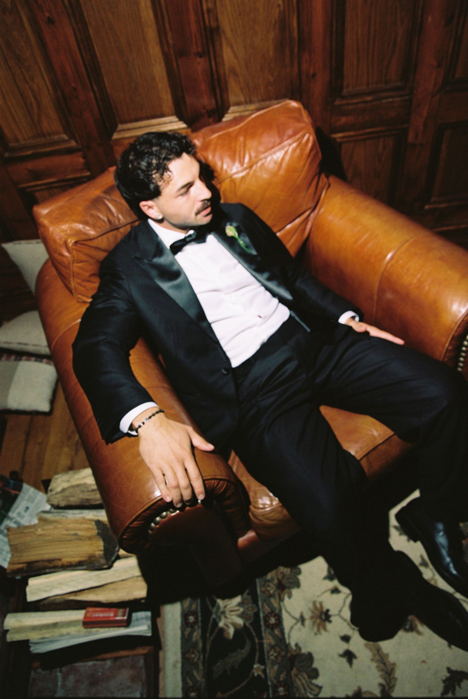 Groom in black tuxedo relaxes on a brown leather sofa surrounded by vintage books in a wood-paneled room.