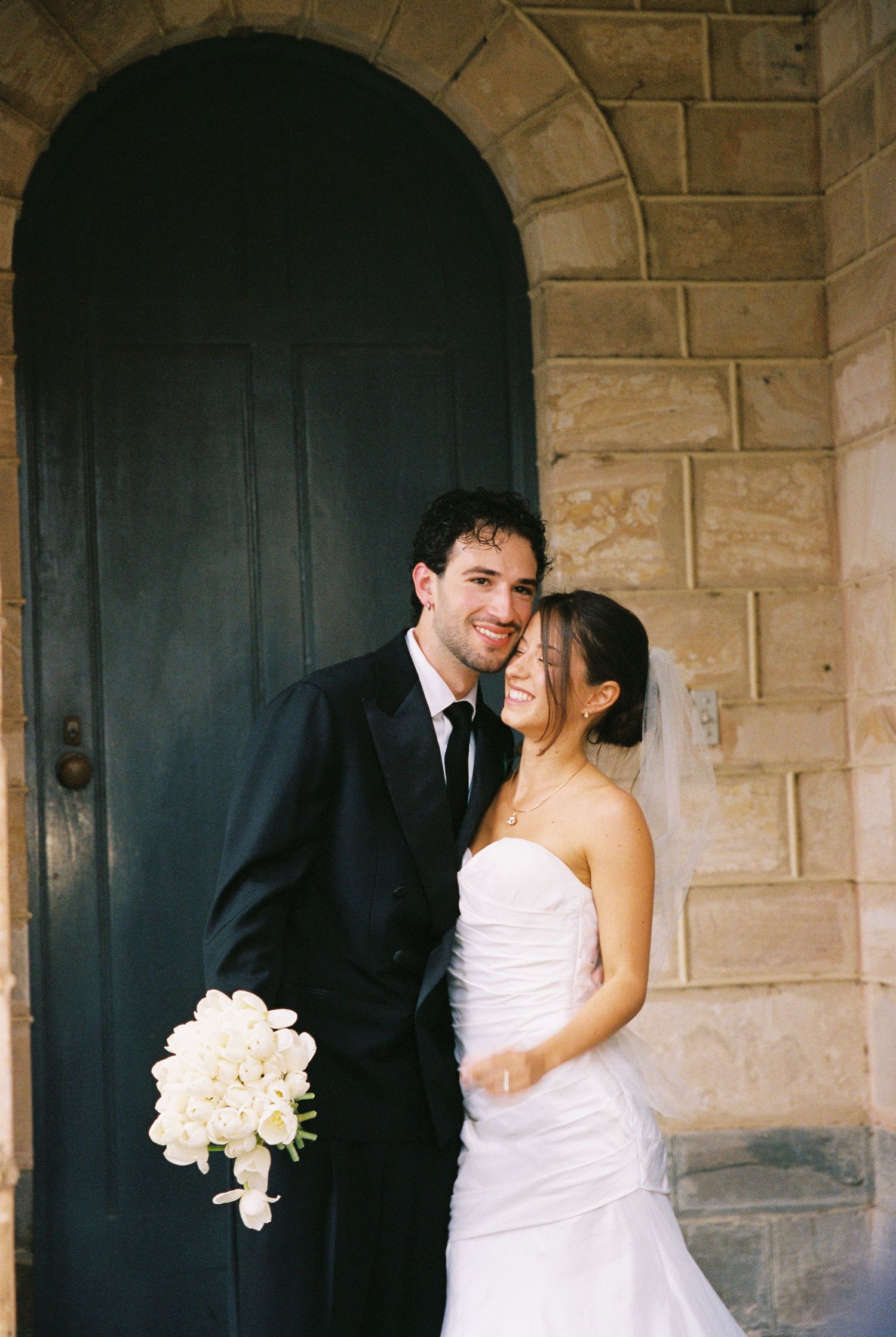 Smiling bride and groom embracing before arched stone doorway, groom in black suit holding white calla lily bouquet.