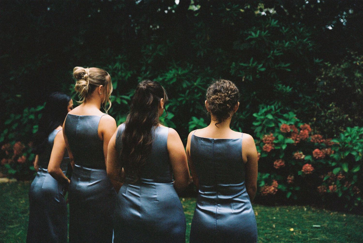 Three bridesmaids in steel blue satin dresses viewed from behind in a garden setting.
