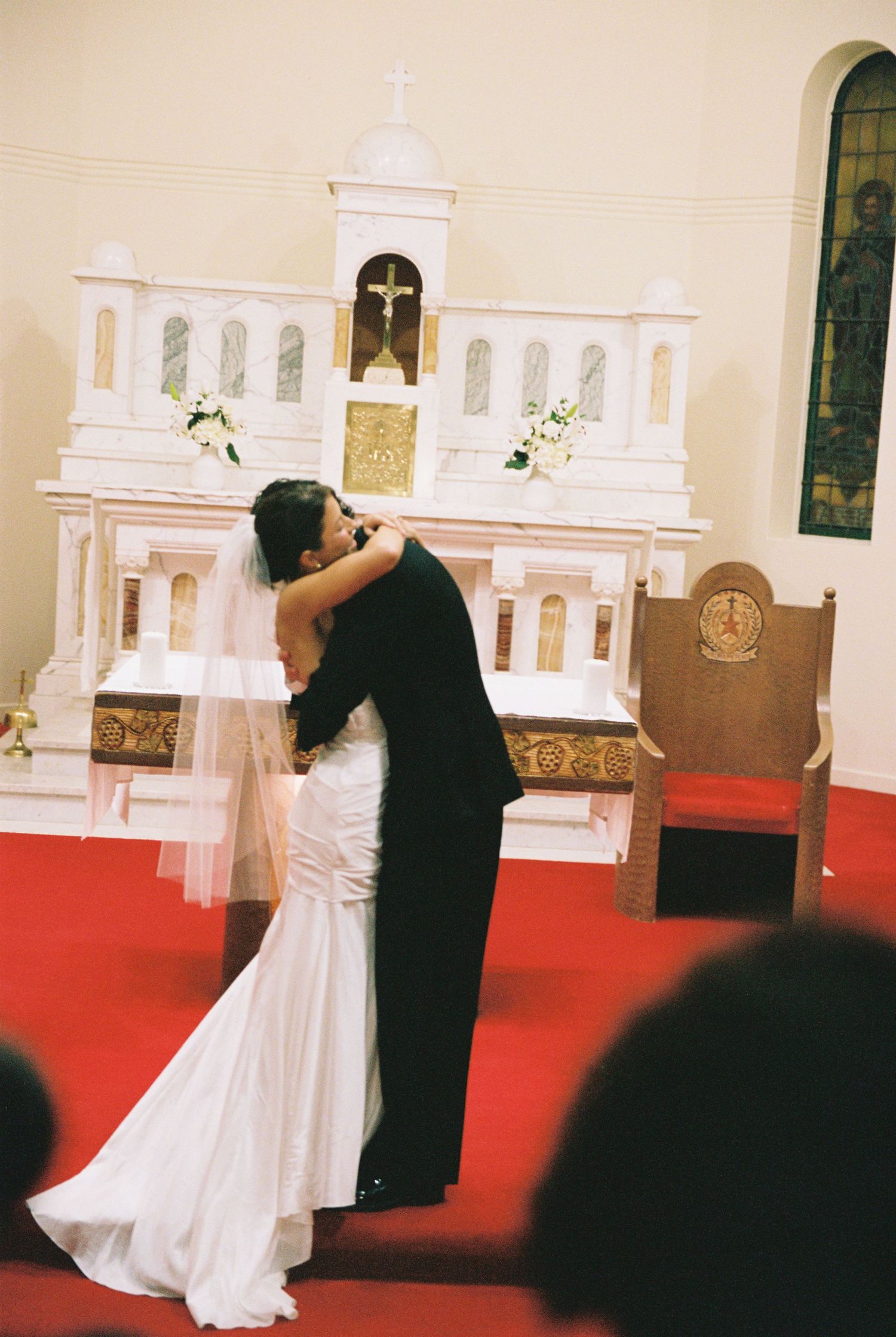 Bride and groom share first kiss at ornate white church altar with red carpet aisle.