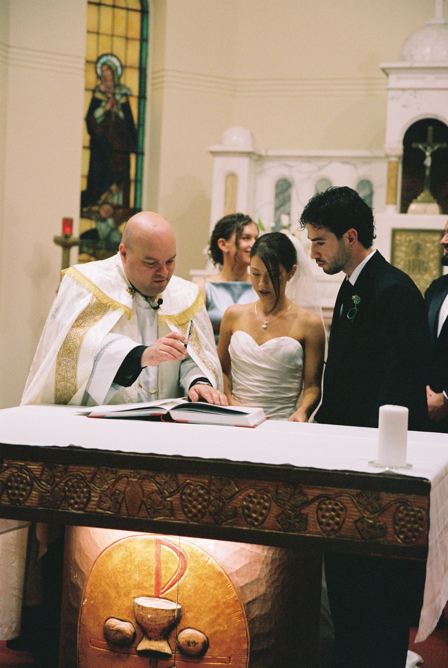 Priest in white vestments signs wedding register with bride and groom at church altar.